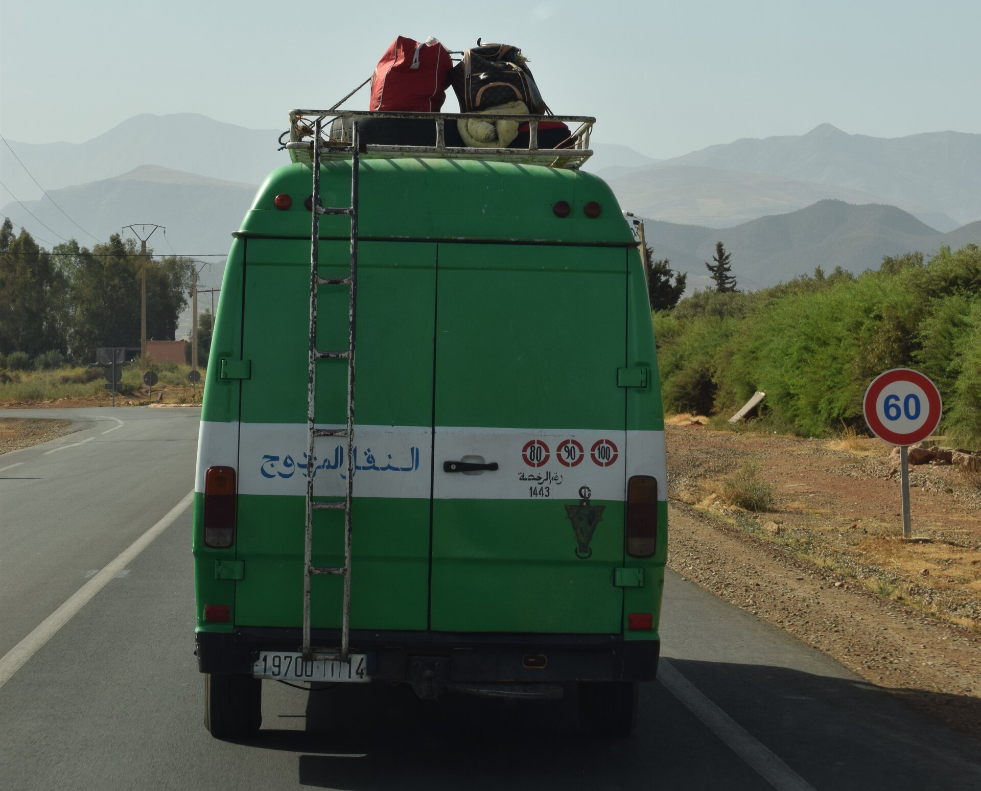 Zdjęcie przedstawia tył zielonego busa, jadącego asfaltową drogą. Na dachu samochodu znajduje się metalowy bagażnik, na którym są widoczne torby i plecaki przywiązane sznurkiem. Po lewej stronie tyłu samochodu widoczna jest wąska drabinka prowadząca do bagażnika na dachu. Po prawej stronie drogi widoczne jest szerokie pobocze oraz pas zieleni. W tle widoczne są pasma górskie.
