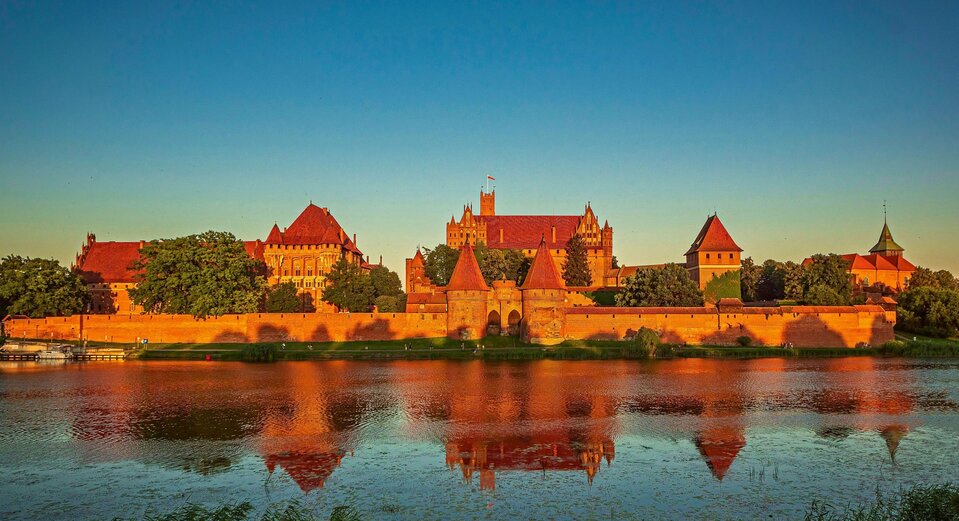 Zdjęcie przedstawia panoramę Zamku Krzyżackiego w Malborku. Zamek jest rozległy, otoczony murem. Stoi nad rzeką. 