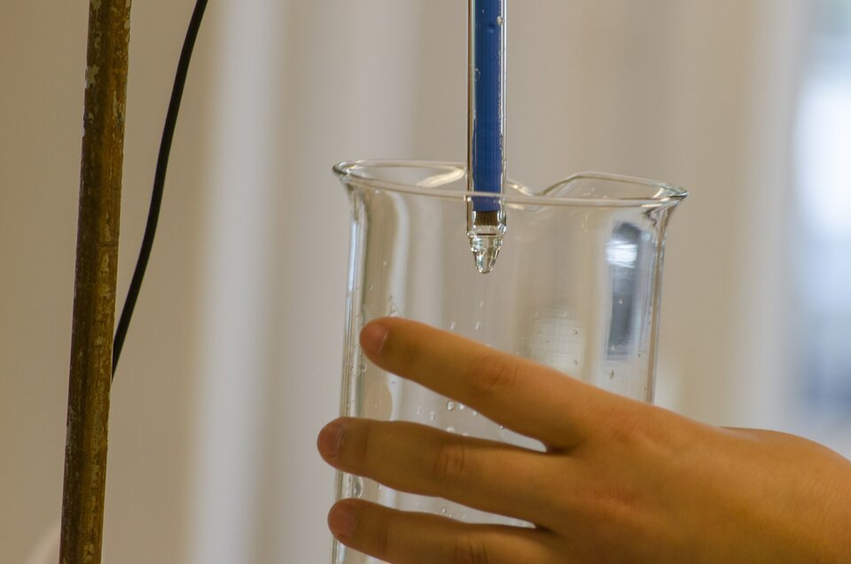 Zdjęcie przedstawia rękę, która trzyma szklany pojemnik. Do pojemnika wlewa się substancja z podłużnej aparatury.