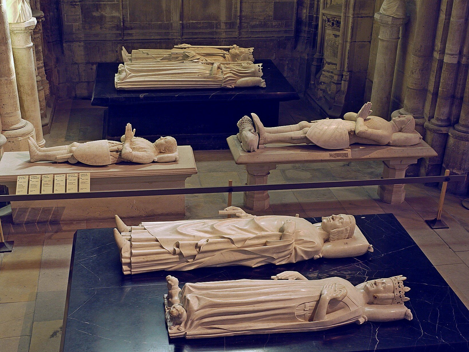 Widocznych sześć nagrobnych figur z nagrobków królewskich, przedstawiających sylwetki męskie i żeńskie w pozycjach leżących. Wszystkie figury zostały wykonane z jasnego kamienia, oszlifowane, przedstawiają elementy anatomiczne ciała i twarzy osób, którym są poświęcone. Dwie figury na pierwszym planie ukazane są w strojach władczyń. Lewa dłoń obu figur położona jest na wysokości klatki piersiowej. Dwie kolejne figury, widoczne w centrum fotografii, mają dłonie złączone na wysokości klatki piersiowej. Dwie figury znajdujące się na dalszym planie, w głębi pomieszczenia, ukazane są w strojach królewskich. Pierwsza z nich trzyma lewą dłoń na wysokości klatki piersiowej, druga zaś - leżąca jako ostatnia -  w prawej dłoni trzyma berło. Położone są na płytach i podestach, po lewej stronie ilustracji znajdują się sygnatury na poręczy, informujące o konkretnym zabytku. Zdjęcie współczesne. 