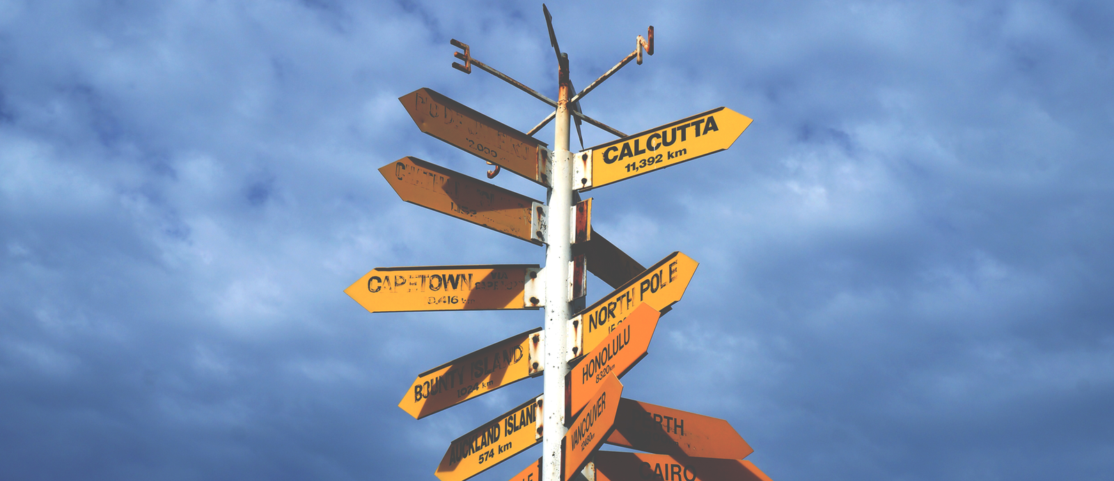 Zdjęcie przedstawia na tle zachmurzonego nieba żółte drogowskazy przyczepione do jednego, białego słupa. Na tabliczkach można odczytać: Calcutta 11392 km, North Pole, Vancouver, Cape Town, Honolulu 8320 km, Auckland Island 574 km, Bounty Island 1024 km, Cairo. Część tabliczek jest nieczytelna.