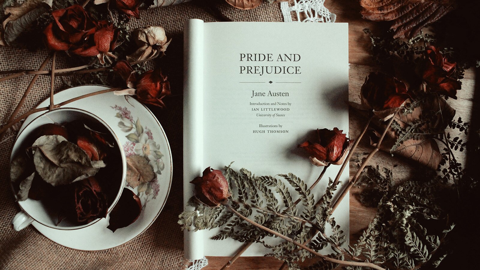 Zdjęcie przedstawia książkę otwartą na stronie tytułowej, gdzie napis głosi "PRIDE AND PREJUDICE" "JANE AUSTEN INTRODUCTION AND NOTES BY IAN LITTLEWOOD. UNIVERSITY OF SUSSEX. ILLUSTRATIONS BY HUGH THOMSON." Obok książki, na spodku, stoi filiżanka wypełniona ususzonymi liśćmi i kwiatami. Wokół książki, jak i na niej, ułożono mnóstwo ususzonych róż i paproci.