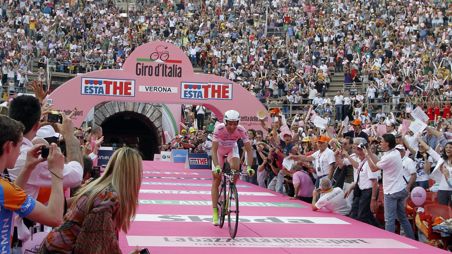 Zdjęcie przedstawia kolarza na rowerze w koszulce lidera wyścigu. Kolarz wjeżdża na stadion wśród tłumu kibiców.