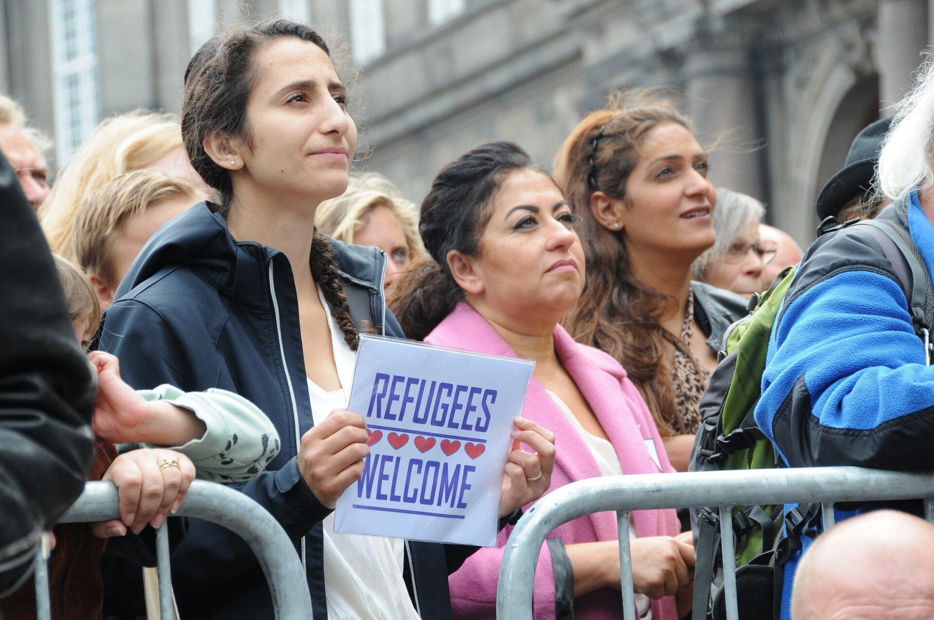 Fotografia przedstawia tłum protestujących pod parlamentem w Kopenhadze. Kobieta na pierwszym planie trzyma w rękach kartkę z napisem: Refugees Welcome.