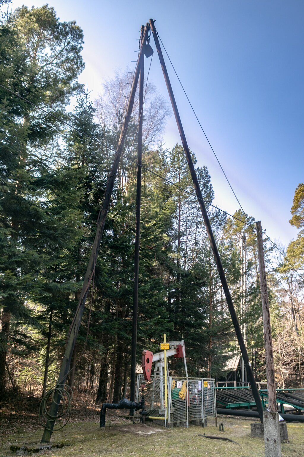 Na zdjęciu znajduje się głowica eksploatacyjna odwiertu ropno gazowego z indywidualnym żurawiem pompowym i trójnogiem do obróbki odwiertu. Głowica eksploatacyjna to urządzenie montowane na powierzchni w celu zamknięcia odwiertu. Żuraw pompowy to inaczej kiwon czyli pompa służąca do pompowania ropy naftowej ze złoża.Najbardziej widocznym elementem na zdjęciu jest trójnóg składający się z trzech nóg ułożonych w kształt ostrosłupa. Pod trójnogiem znajduje się czarna głowica odwiertu, a obok niej kiwon pompowy. Od głowicy eksploatacyjnej odchodzi rura umieszona w ziemi tuż obok. Głowica połączona jest z łbem wahacza za pomocą linek zwanych żerdzią dławikową. Łeb wahacza jest koloru czerwonego i znajduje się w lewej części ramienia kiwona zwanego wahaczem. Łeb jest w kształcie głowy młotka. Po drugiej stronie wahacza znajduje się szare połączenie z podciągaczami, które połączone są do obciążników. W środkowej części wahacza znajduje się szara rama nośna. Za ramą znajduje się prostokątna skrzynia oraz silnik kiwona. Za kiwonem znajdują się konstrukcje przypominające panele słoneczne lub rampy do prac obróbczych. W tle znajdują się drzewa iglaste.
