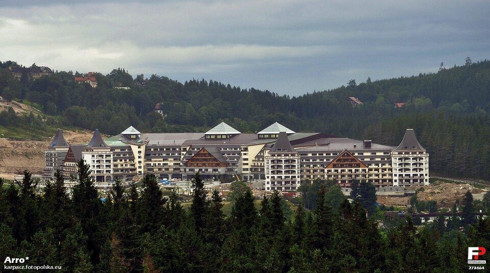 Na zdjęciu znajduje się wielki, rozległy hotel. Składa się z kilku połączonych ze sobą skrzydeł. Na końcach skrzydeł znajdują się spiczaste dachy. Budynek główny i oba skrzydła mają trójkątne portyki. teren przed hotelem jest zasłonięty przez znajdujące się na pierwszym planie świerki. W tle, za hotelem, są łagodne stoki, porośnięte gęsto drzewami, między którymi widać fragmenty dachów kilku budynków mieszkalnych.