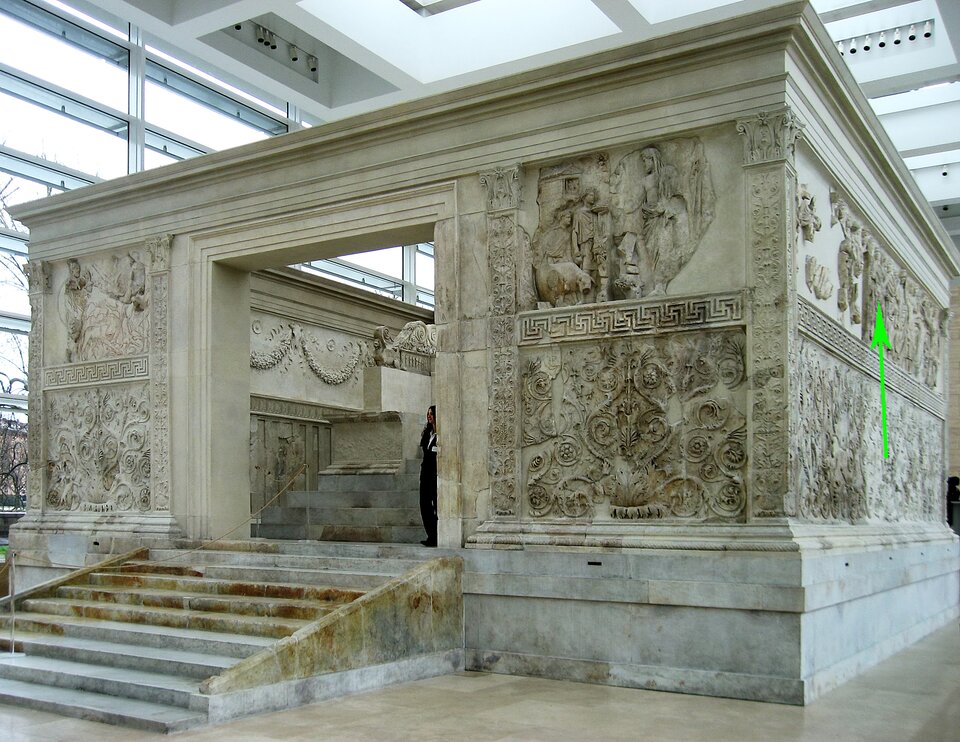 Ołtarz Pokoju Augusta, I w p.n.e., Muzeum Ara Pacis Augustae, Rzym, Włochy Na fotografii znajduje się duża, kamienna budowla ozdobiona licznymi płaskorzeźbami i dekoracyjnymi ornamentami. Bogato rzeźbione reliefy przedstawiają procesje, postacie mitologiczne oraz motywy roślinne. W górnej części prawej ściany budowli na fotografii, zielona strzałka skierowane wektorem w dół, wskazuje wyrzeźbione postaci idące w procesji. Płaskorzeźba zachowana jest fragmentarycznie. Obiekt znajduje się wewnątrz przestronnej, nowoczesnej sali muzealnej z wysokimi oknami i jasnym sufitem. Do wnętrza budowli prowadzą szerokie marmurowe schody.