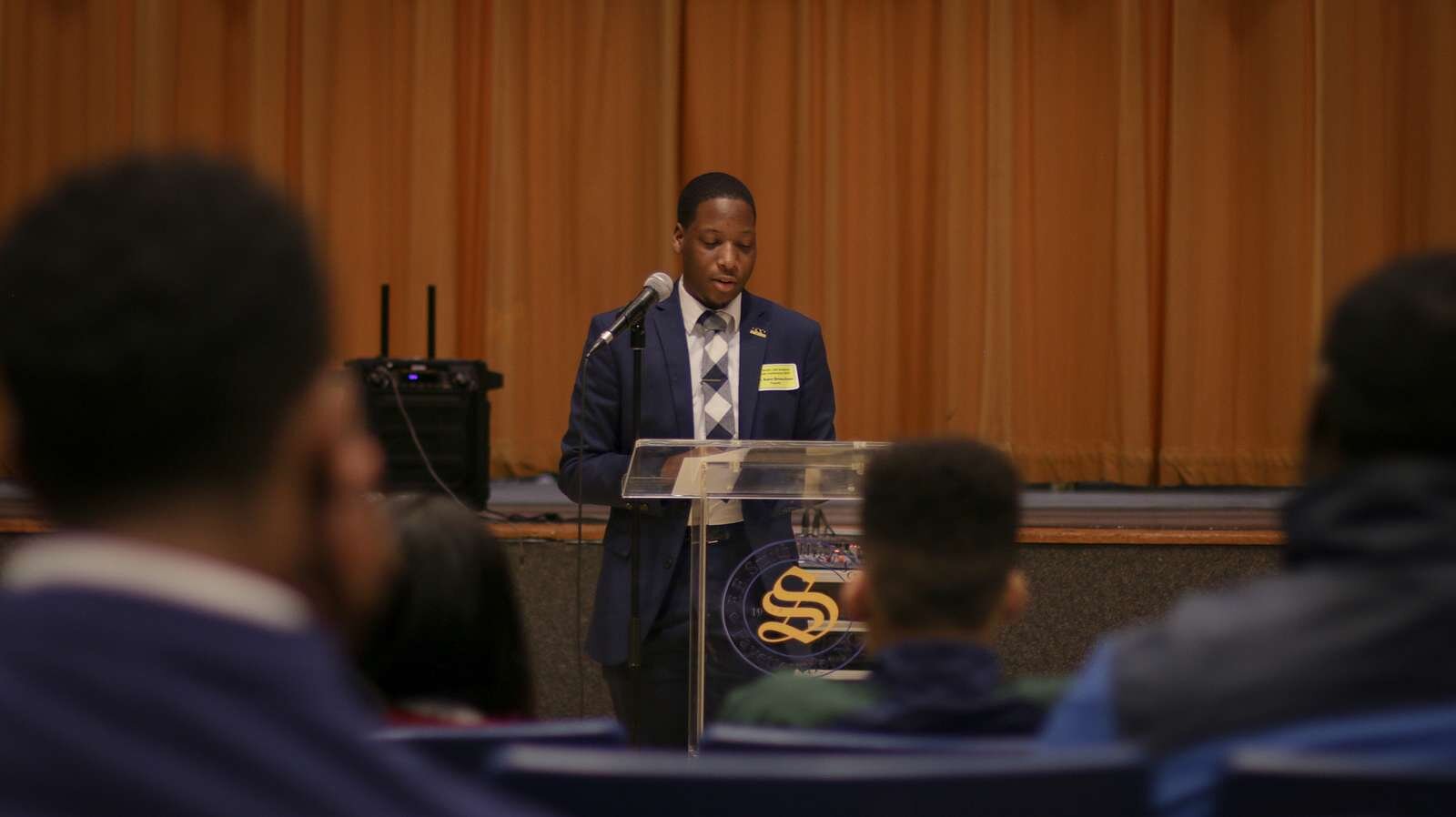 Zdjęcie przedstawiające stojącego przy mównicy mężczyznę, który spogląda na kartkę leżącą na pulpicie. Widoczne są też głowy osób siedzących i słuchających wystąpienia.