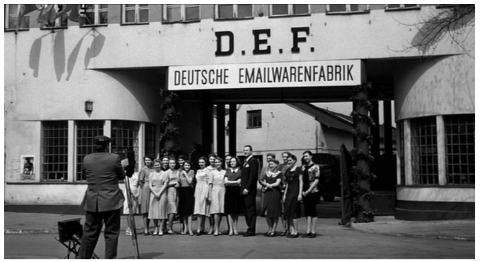 Zdjęcie przedstawia duży budynek z niemieckim szyldem D. E. F. Deutsche Emailwarenfabrik. Przed wejście stroi grupa kobiet w letnich sukienkach, a pomiędzy nimi Schindler w garniturze. Naprzeciw nich stoi fotograf robiący im zdjęcia.