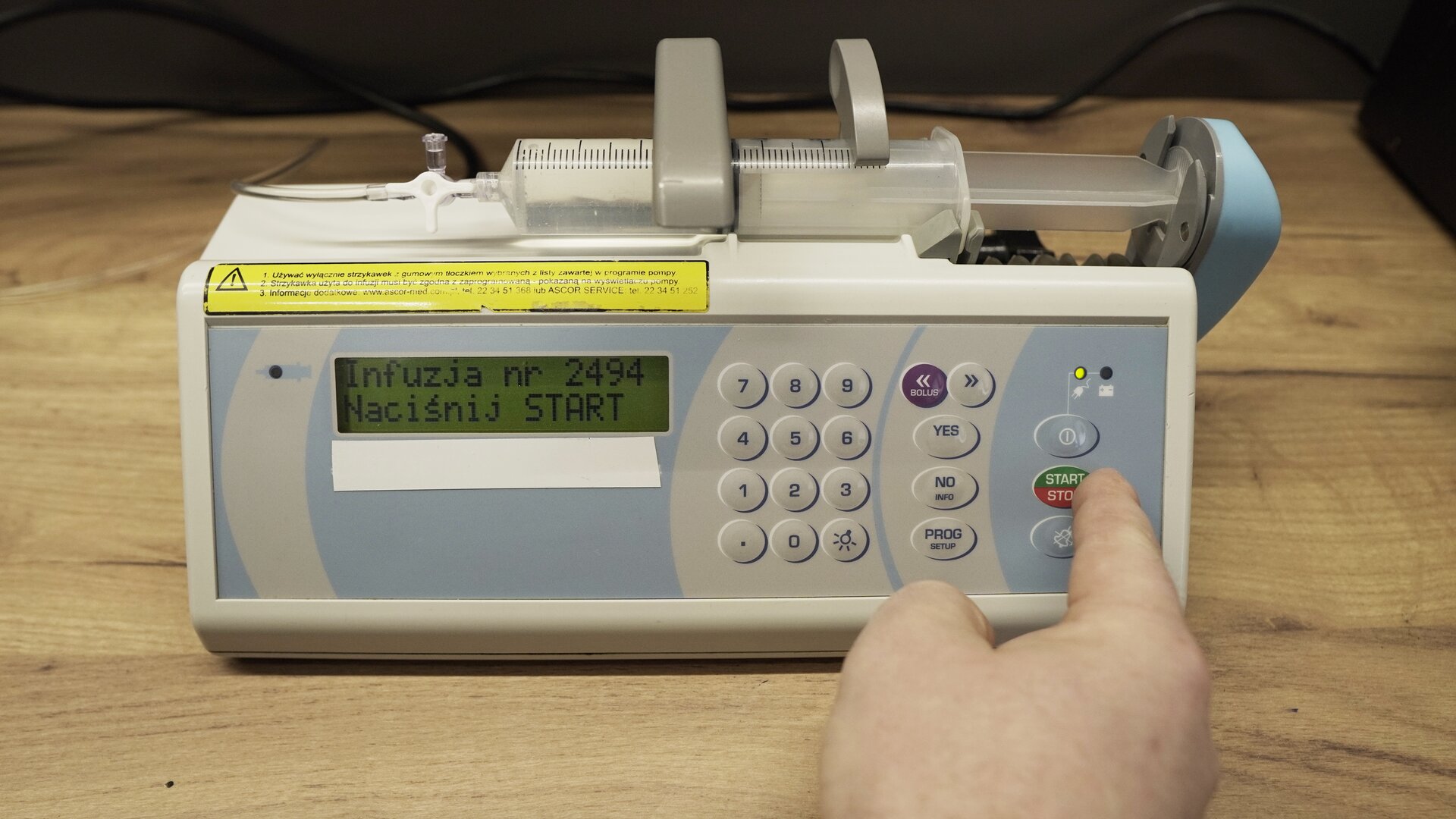 Na zdjęciu strzykawka wypełniona wodą znajduje się w ramieniu pompy infuzyjnej. Na ekranie aparatu wyświetlana jest informacja: "Infuzja 2494 Naciśnij przycisk start". Technik wciska przycisk star/stop na obudowie aparatu.