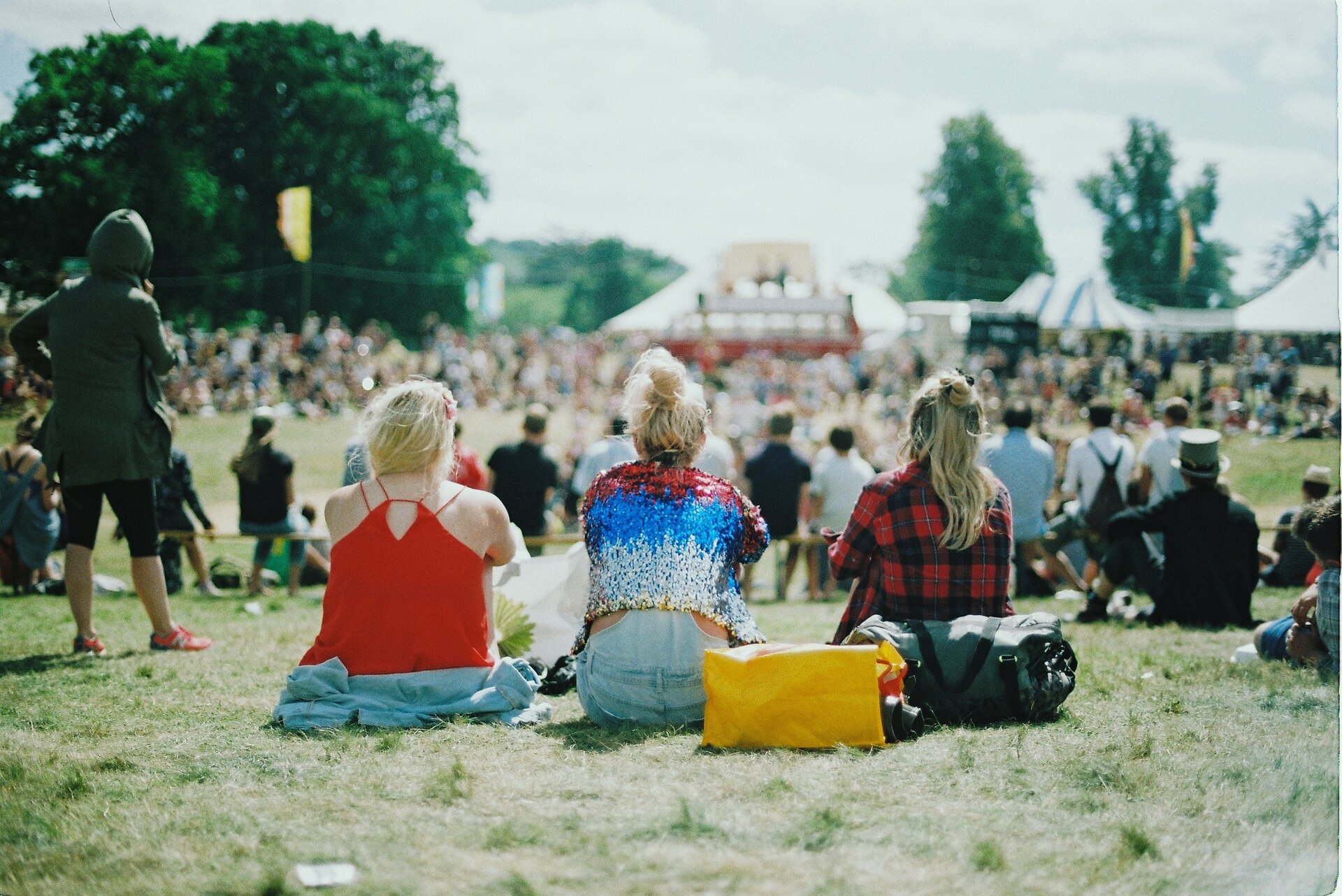 Zdjęcie przedstawia młodzież siedzącą na trawie podczas festiwalu. W tle są tłumnie zgromadzeni ludzie, scena oraz namioty.