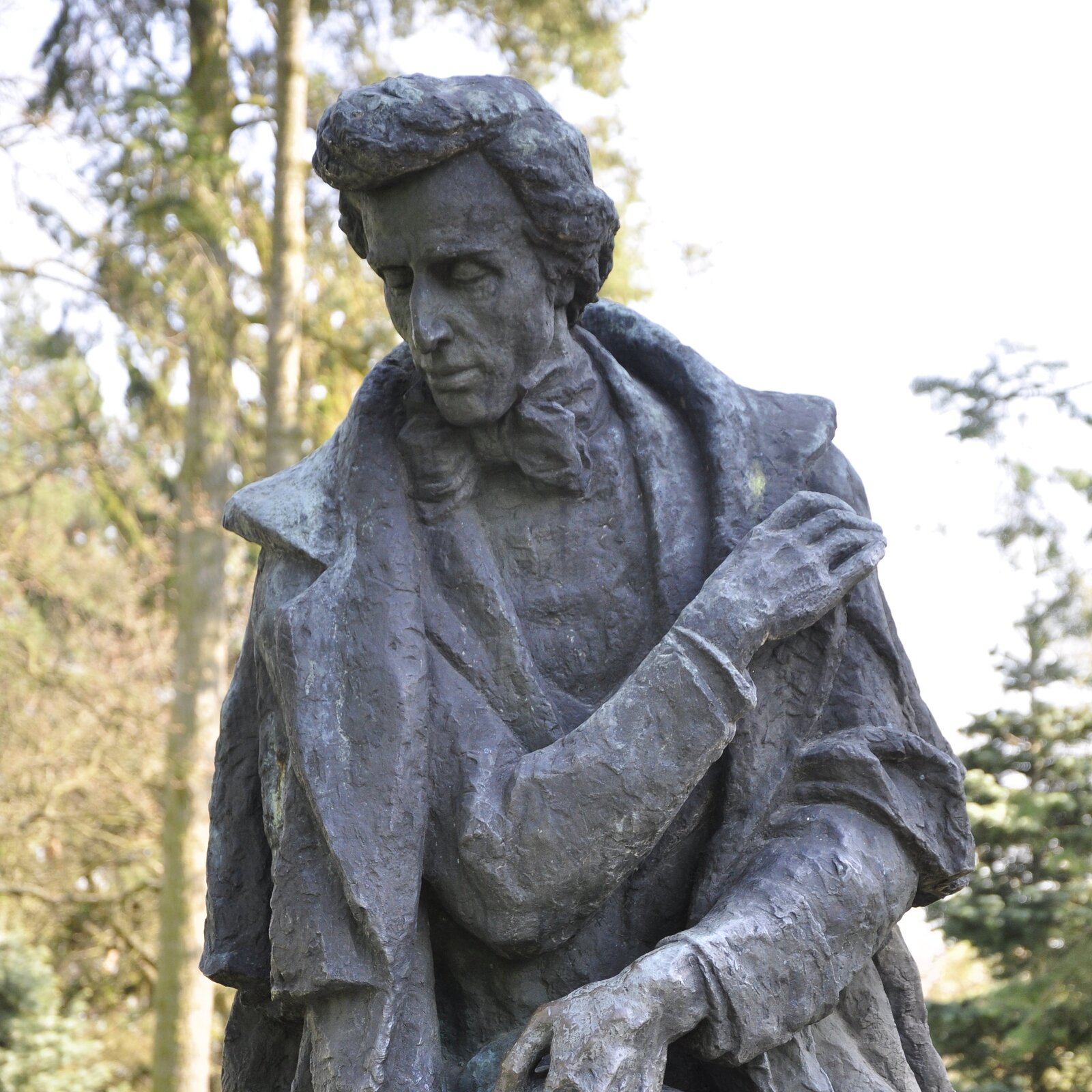 Zdjęcie przedstawia fragment pomnika Fryderyka Chopina. Mężczyzna prawą dłonią przytrzymuje lewą część płaszcza, a lewą rękę ma założoną z prawej strony. Mężczyzna nosi marynarkę oraz dużą kokardę na szyi. Głowę ma schyloną w dół, usta zamknięte, oczy przymknięte.