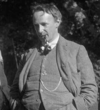 Zdjęcie przedstawia dojrzałego mężczyznę. Ma krótkie, lekko rozczochrane włosy. Ma w ustach fajkę. Trzyma ręce w kieszeniach spodni.