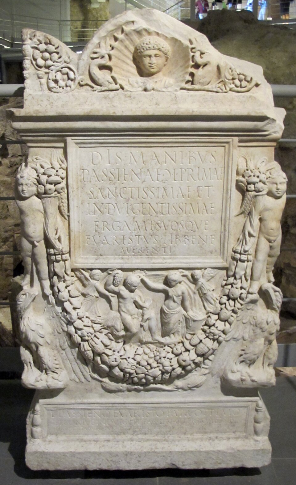 Ilustracja przedstawia Ara Funeraria Luciusa Aufidiusa Aprilisa, ołtarz grobowy z grobowca VI nekropolii przy Via Triumfalis, datowany na ok. 60–70 n.e., przechowywany w Watykanie. Ukazuje frontową stronę marmurowego ołtarza, bogato dekorowaną reliefami. Głównym motywem są obszerne girlandy z liści, owoców i kwiatów, rozpięte szeroko w poprzek kompozycji. Girlandy podwieszone są na głowach Jowisza Ammona o charakterystycznych baranich rogach oraz na głowach baranów, umieszczonych na narożach ołtarza. Poniżej girland widoczne są drobne, misternie opracowane postacie, wśród nich tańczące kobiety, ukazane w lekkim ruchu, z uniesionymi ramionami i rozwianymi draperiami. W dekoracji pojawiają się także orły, przedstawione z rozpostartymi skrzydłami, umieszczone symetrycznie po bokach. Centralną część kompozycji zajmuje prostokątna tablica inskrypcyjna z równymi, głęboko rytymi literami.
