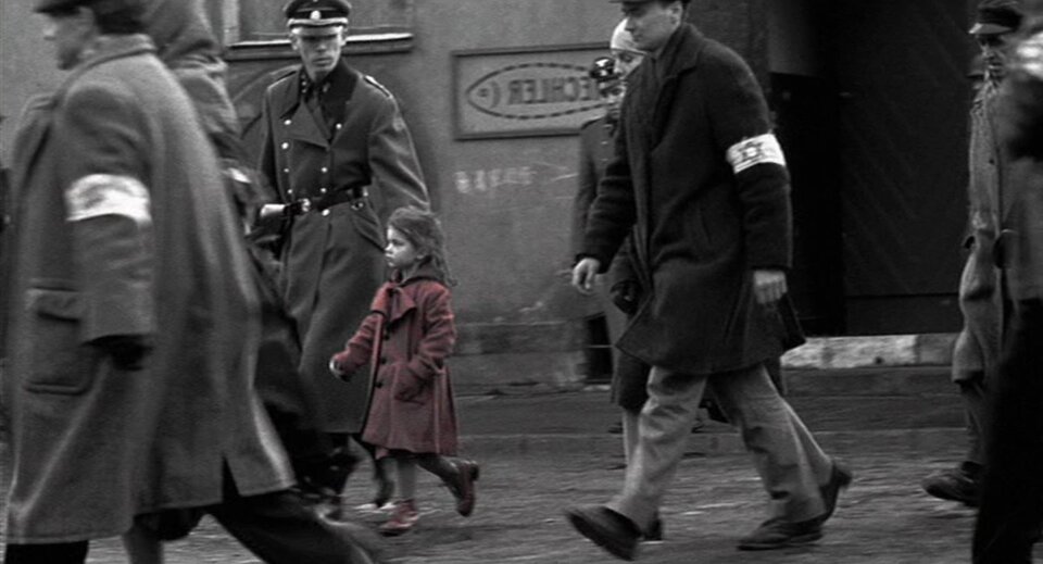 Zdjęcie czarno‑białe przedstawia ulicę. Idą nią ludzie z opaskami z gwiazdą Dawida eskortowani przez Niemców. Pomiędzy nimi idzie dziewczynka w czerwonym płaszczyku.
