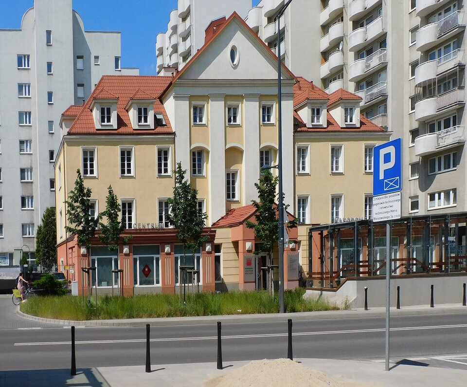 Fotografia przedstawia gmach o klasycznej bryle w tonacji biało‑beżowej. Prawa strona przed wejściem zabudowana szklanym tarasem. Za budynkiem i po jego prawej stronie widoczna współczesna zabudowa mieszkalna. Przed budynkiem droga i małe drzewa.