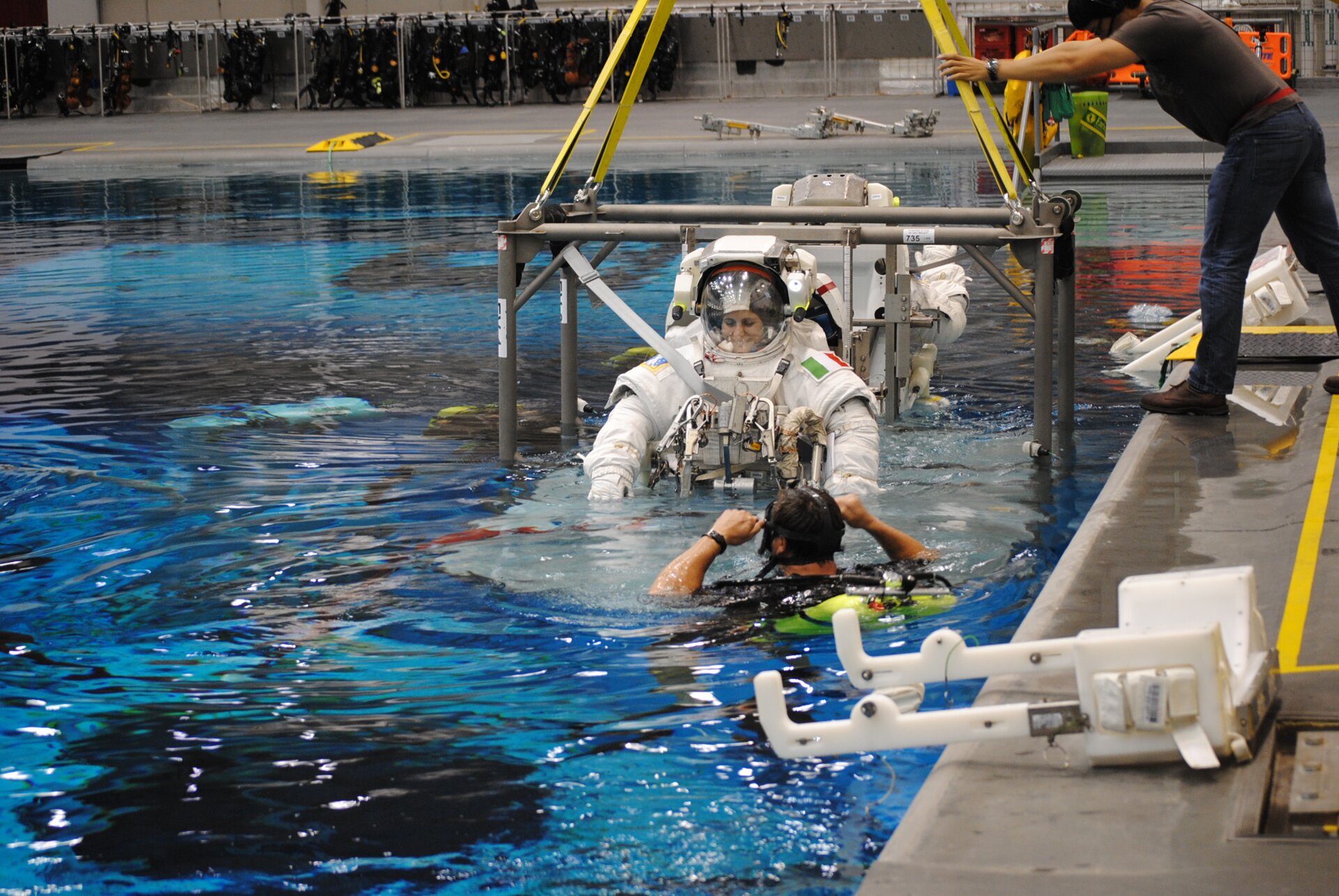 Zdjęcie przedstawia astronautkę podczas szkolenia. Astronautka ubrana jest w skafander. Ćwiczenia odbywają się w basenie. Astronautka zanurzona jest do połowy. W basenie znajduje się również mężczyzna. Mężczyzna wyposażony jest w sprzęto do nurkowania.