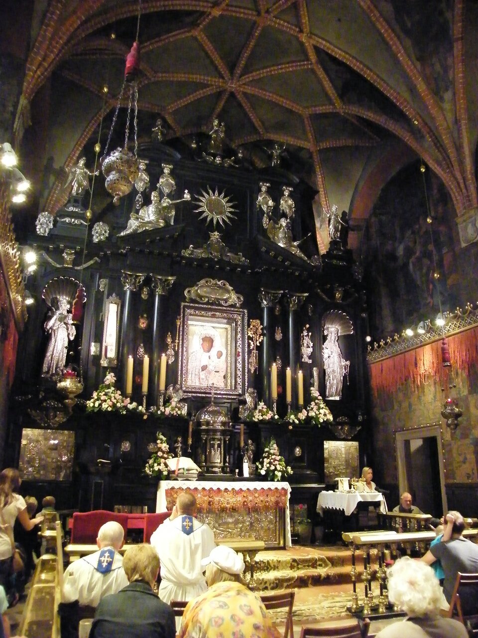 Zdjęcie przedstawia wnętrze kaplicy Matki Boskiej Częstochowskiej na Jasnej Górze. W centrum znajduje się obraz, do którego modlą się duchowni i wierni. Widoczne są złote ornamenty, posagi świętych i aniołów, ołtarz jest wykonany z drewna pomalowanego na czarno. Na ścianach widnieją freski i obrazy. 