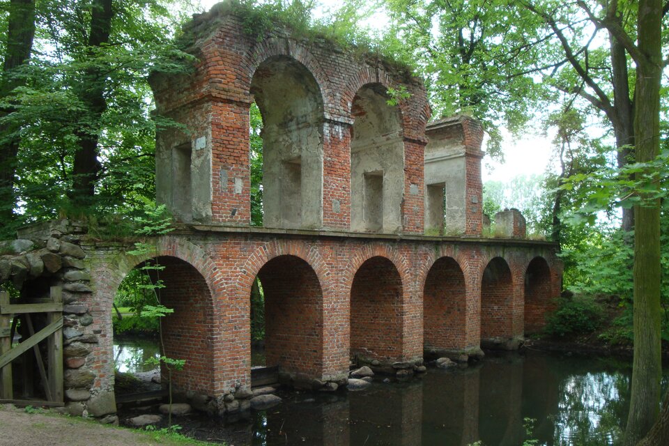 Ilustracja przedstawia akwedukt w parku w Arkadii w Nieborowie, wzorowany na architekturze wodociągów rzymskich. Budowla jest wzniesiona nad kaskadą wodną. Jest dwukondygnacyjna, z czerwonej cegły