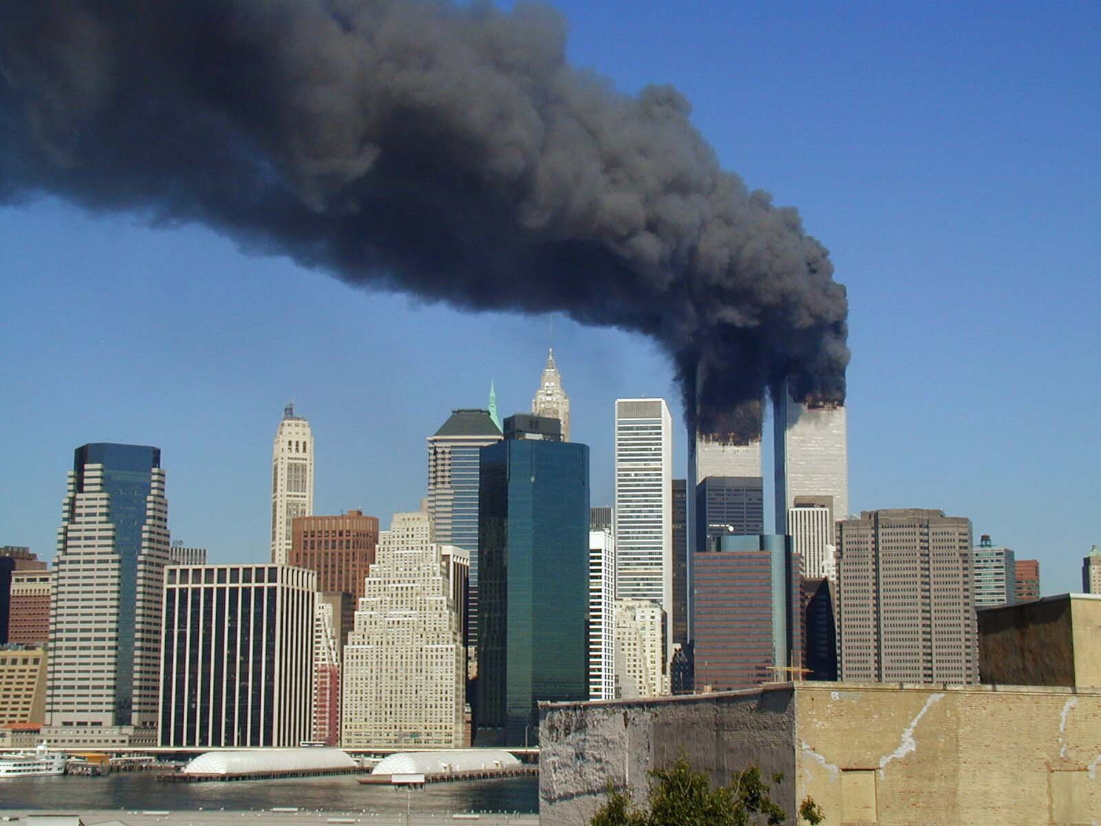 Zdjęcie przedstawia metropolię z wieżowcami. Z górnej części dwóch drapaczy chmur wydobywa się gęsty, czarny dym.   
