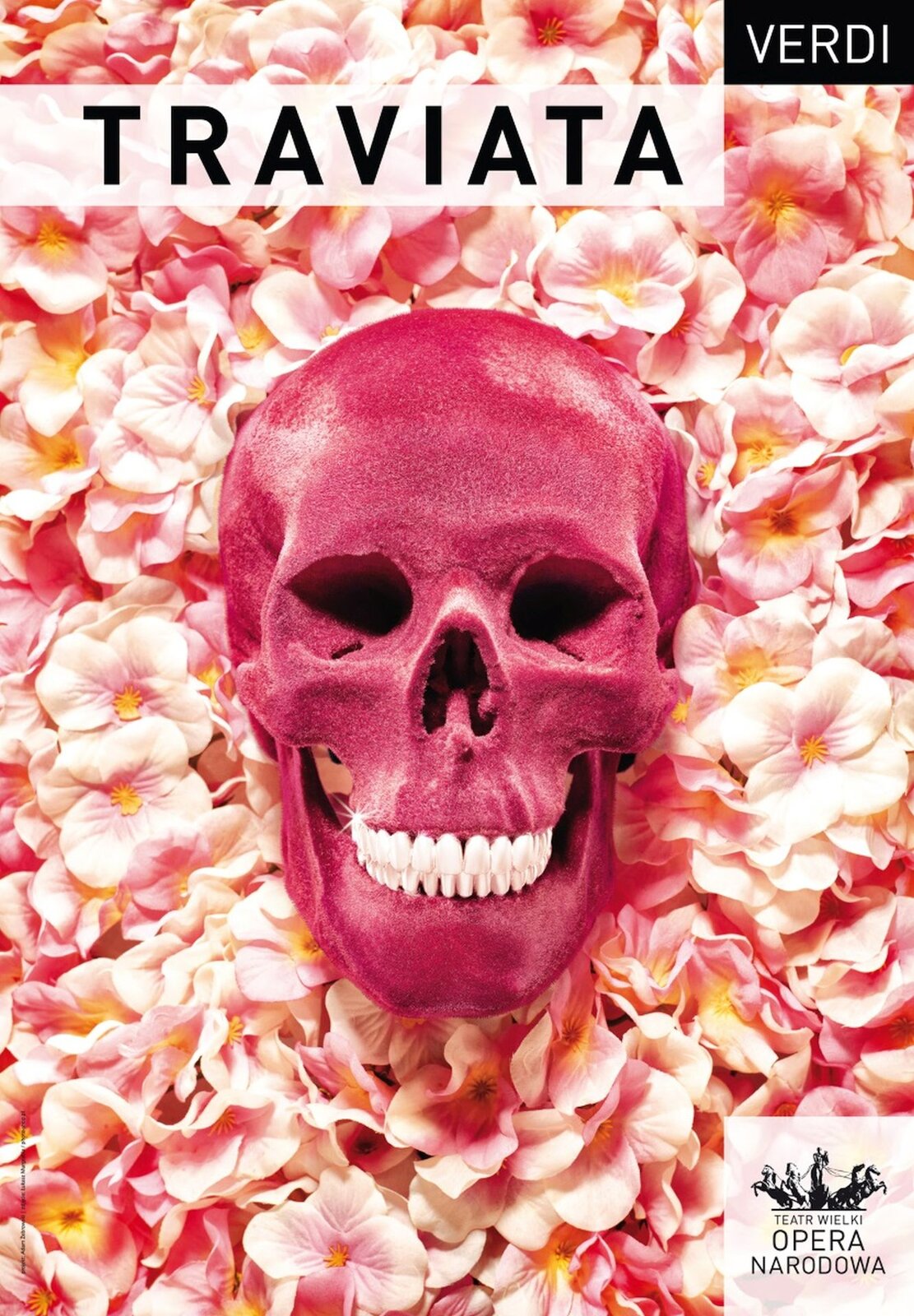 Plakat promujący przedstawienie „La Traviata” Giuseppe Verdiego w reżyserii Mariusza Trelińskiego. Przedstawienie odbyło się w Teatrze Wielkim - Operze Narodowej w Warszawie. Na plakacie na tle kwiatów w pastelowych kolorach, leży czaszka w intensywnym kolorze różowym.