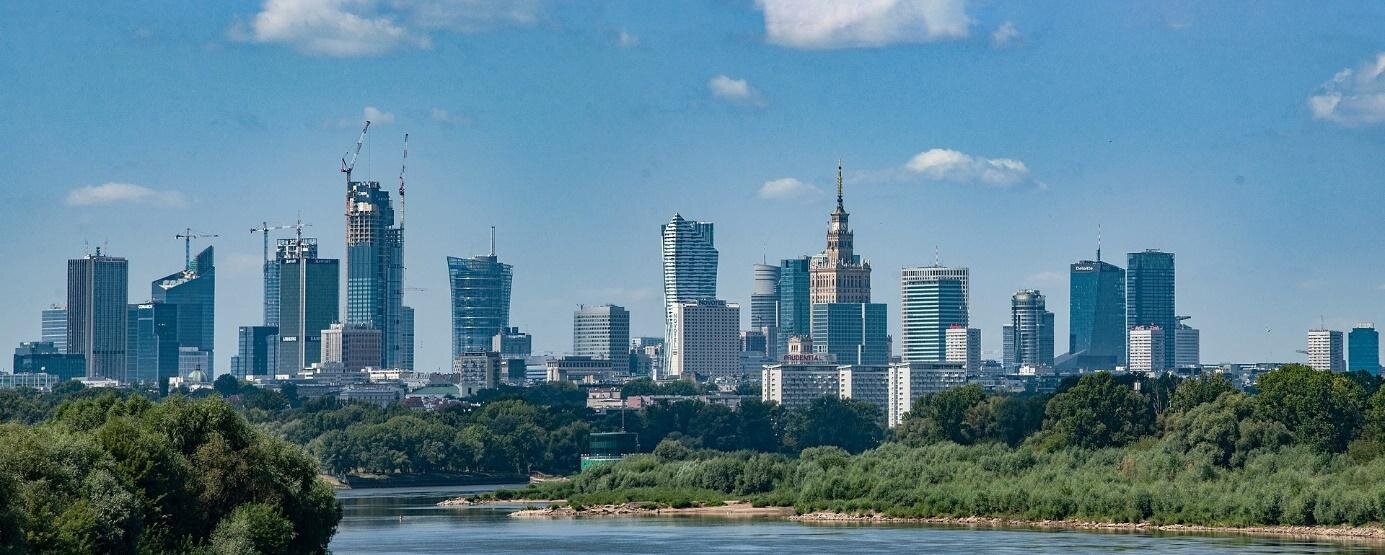 Zdjęcie przedstawia panoramę centrum Warszawy. Na pierwszym planie rzeka, na brzegach której rosną krzewy i drzewa. W oddali biurowce i wieżowce górujące nad miastem. 