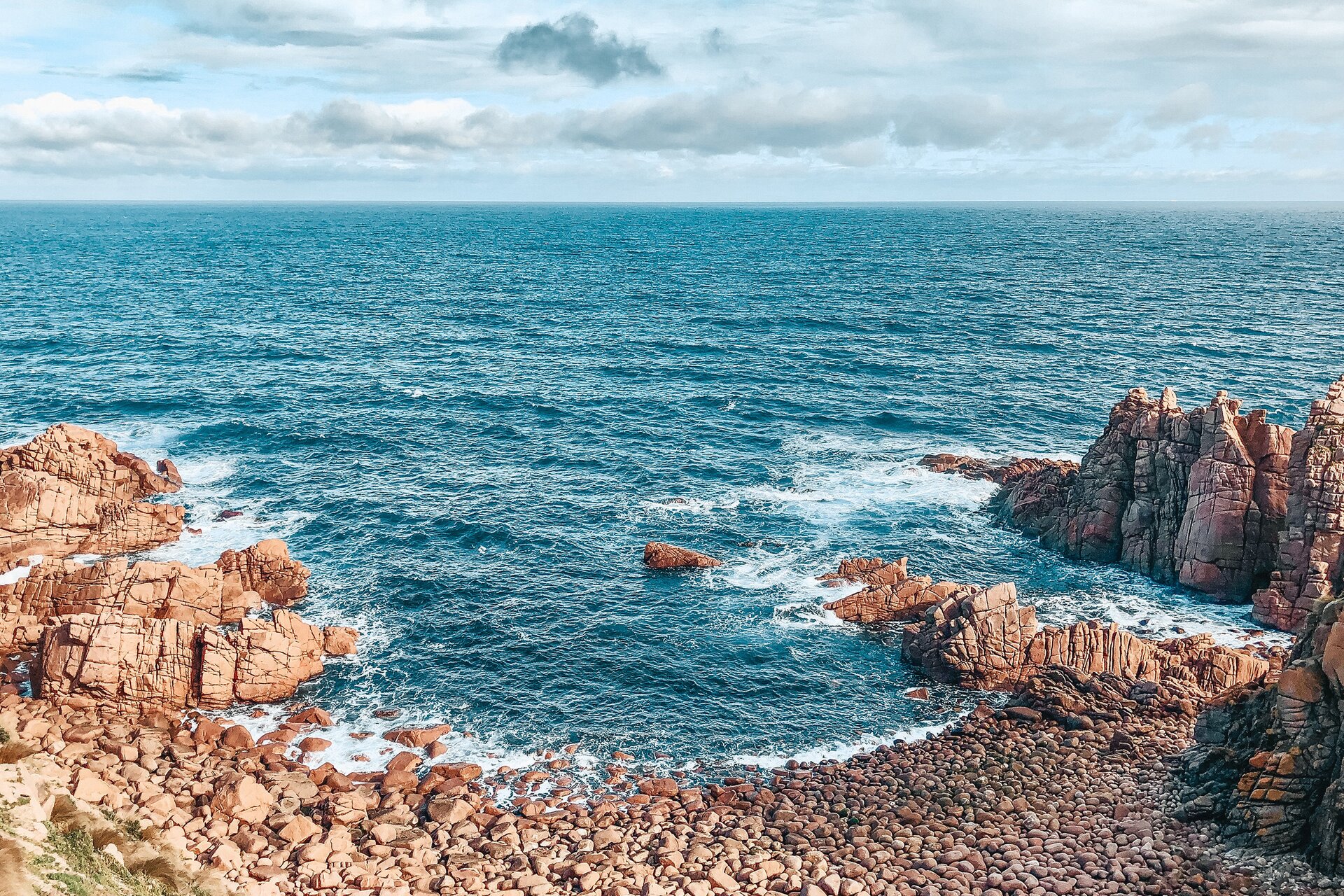 Zdjęcie przedstawia fragment skalistego brzegu oraz zbiornik wodny z niebieską, lekko pofalowaną taflą wody. Tuż przy brzegu widoczne są białe, spienione fale, które uderzają o skały. 
