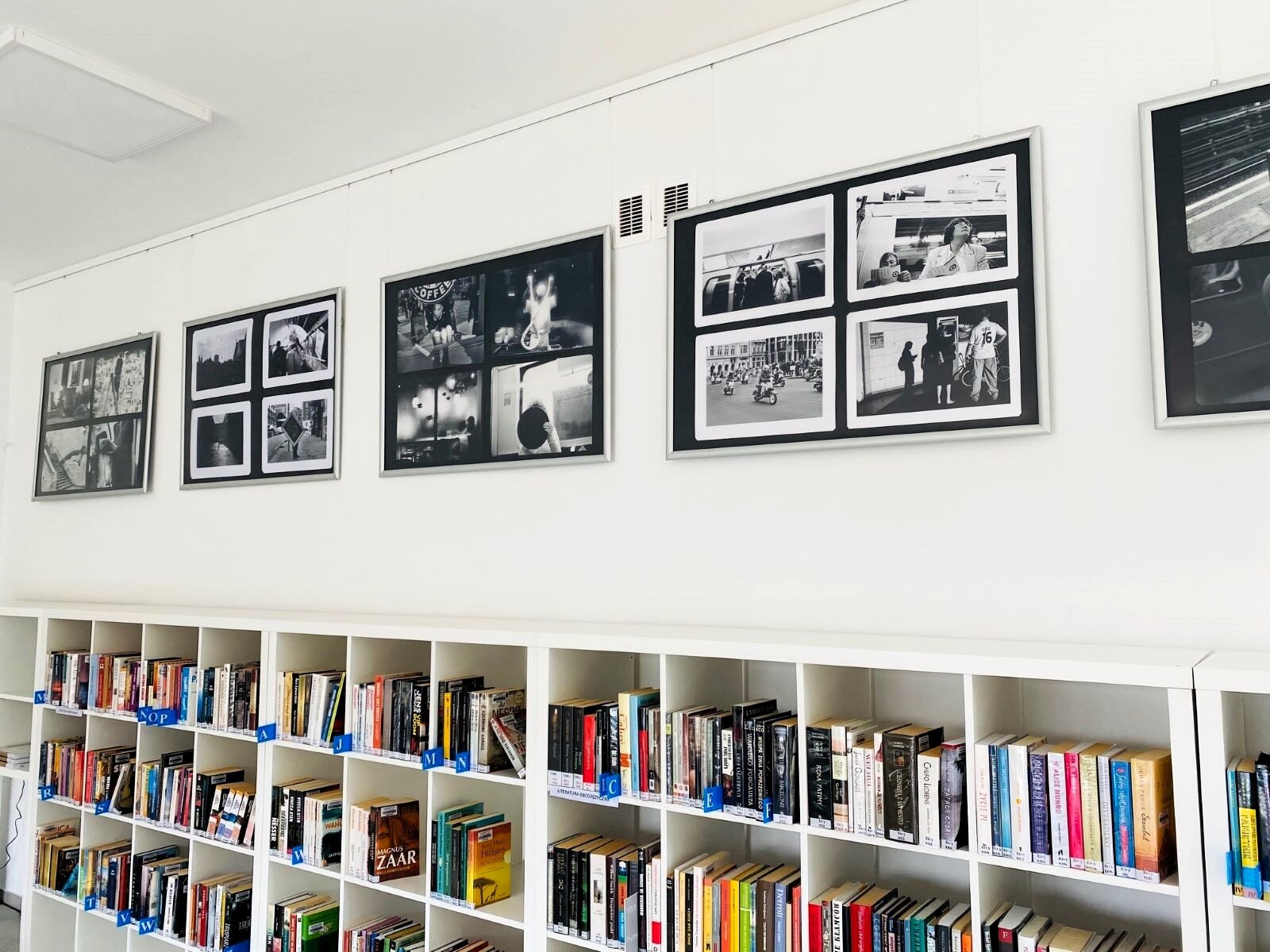Zdjęcie pokazuje wystawę fotografii w bibliotece. Fotografie są  powiększone do dużych rozmiarów, pogrupowane po cztery i oprawione w proste, ciemne ramki, odcinające się od jasnej ściany. Zdjęcia zostały powieszone rzędem, w regularnych odstępach nad bibliotecznymi półkami.