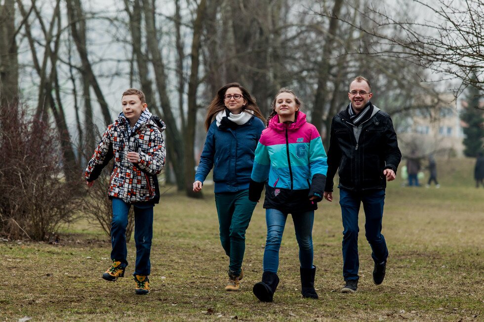 Fotografia przedstawia cztery osoby, które spacerują po parku. Osoby ubrane są w kurtki, szaliki i ciepłe buty. W tle widoczne są bezlistne drzewa.