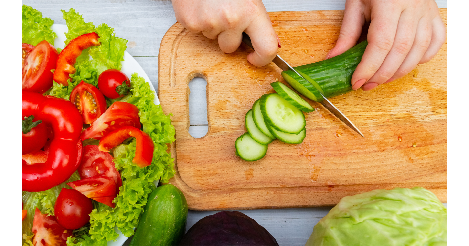 Zdjęcie przedstawia ręce osoby, która kroi na plasterki świeżego ogórka na desce do krojenia. Obok na talerzu znajdują się pokrojone pomidory i papryka, które leżą na listkach sałaty.