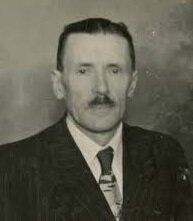 Zdjęcie przedstawia portret starszego mężczyzny. Jego twarz jest pociągła, znajdują się na niej liczne zmarszczki. Mężczyzna ma krótkie, zaczesane ku górze włosy. Jego oczy są głęboko osadzone, nos duży, prosty. Mężczyzna ubrany jest w garnitur, koszulę i krawat.