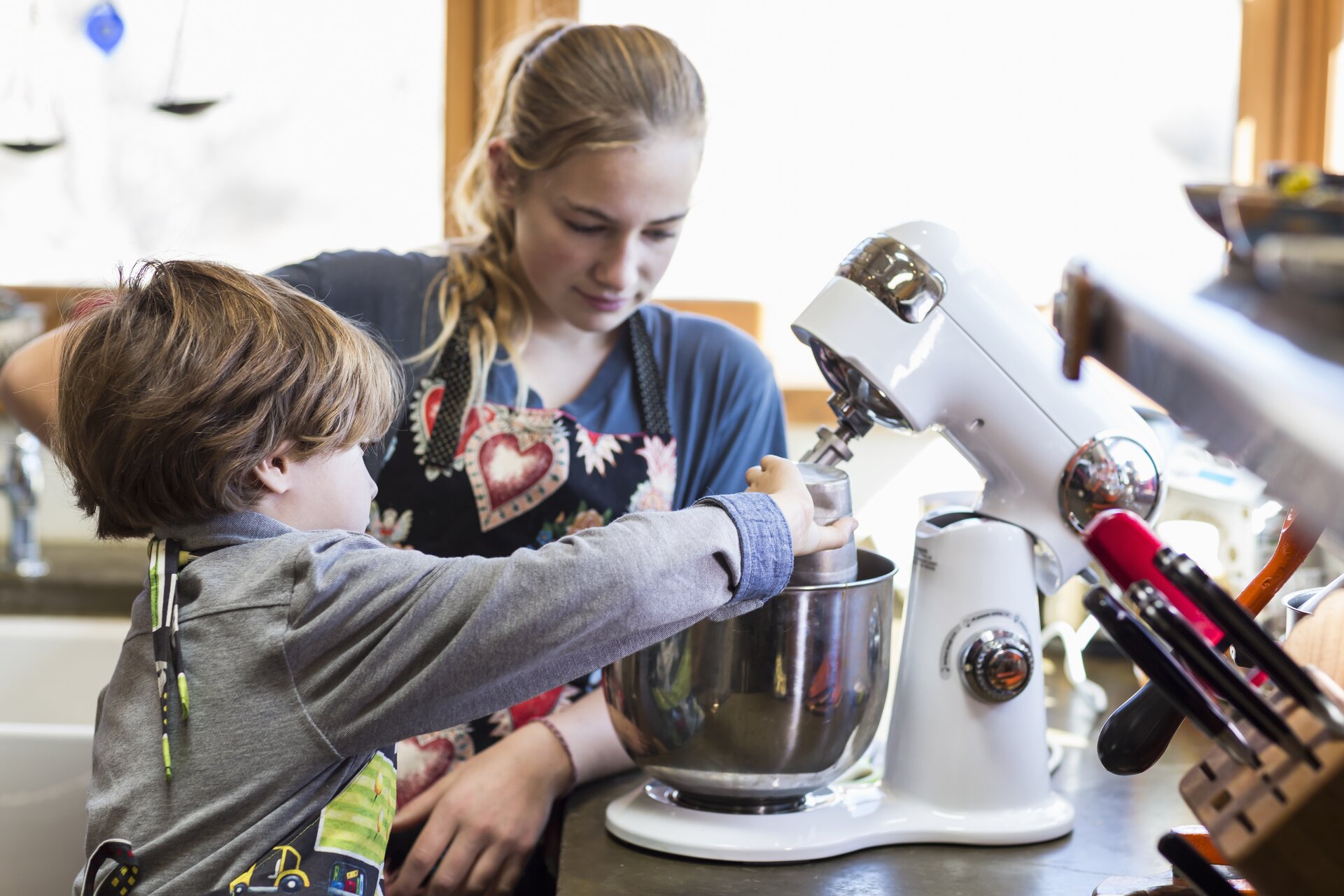 Zdjęcie przedstawia wnętrze kuchni. Na pierwszym planie widoczna jest młoda kobieta i dziecko, którzy stoją przy blacie. Dziecko w prawej ręce trzyma kubek, z którego wysypuje coś do dużego metalowego pojemnika robota kuchennego. Młoda kobieta obserwuje pracę dziecka. Po prawej stronie widoczny jest stojak z nożami.