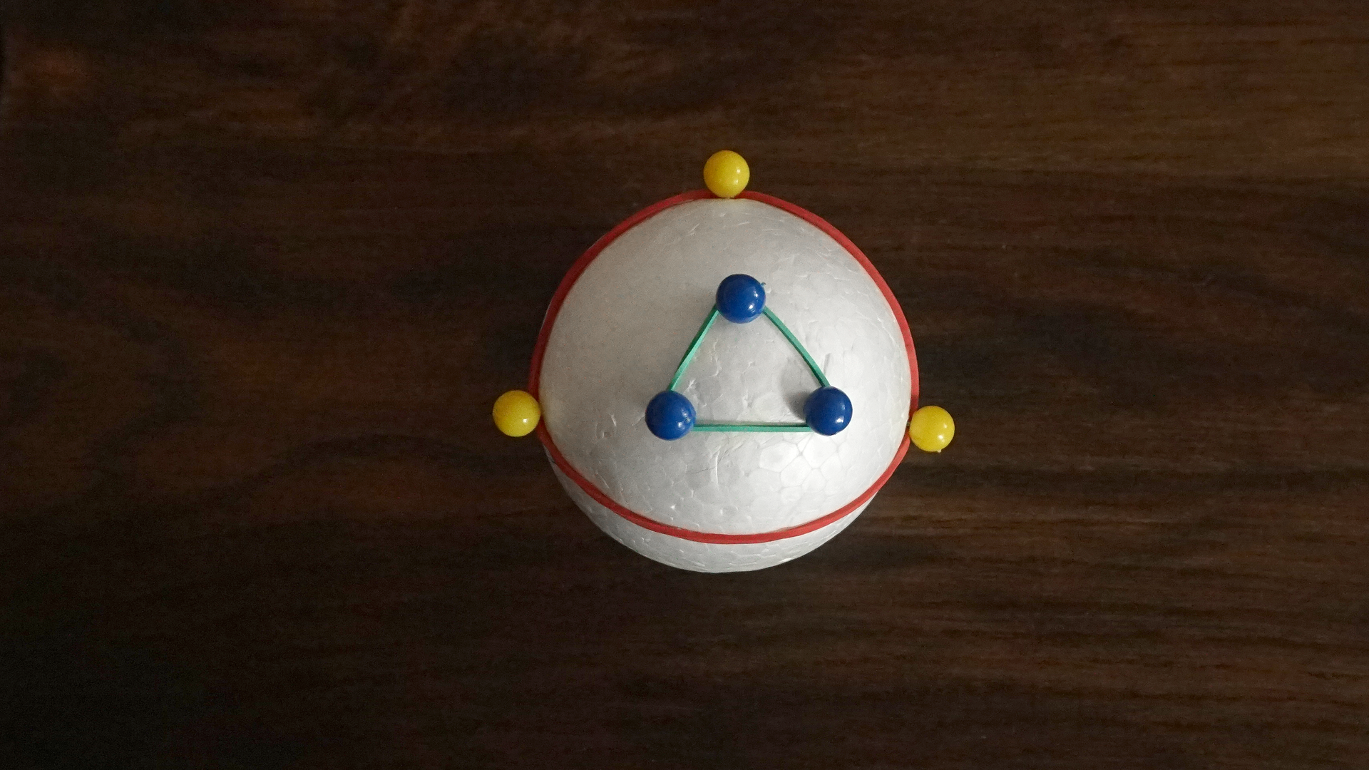 Zdjęcie przedstawia styropianową kulę, w którą wbito przy jednym biegunie trzy szpilki blisko siebie i otoczono je gumką recepturką. Gumka utworzyła trójkąt, którego boki są prawie prostymi łukami. Drugi trójkąt składa się ze szpilek wbitych daleko od siebie w okolicy równika. Kąty w tym trójkącie mają znacznie większe miary, a jego boki są łukami.