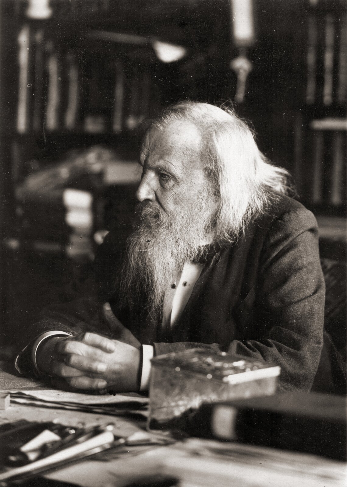 Fotografia czarno-biała przedstawiająca rosyjskiego chemika Dymitra Mendelejewa z okresu późnej starości. Uczony siedzi przy biurku otoczony książkami i spogląda z zamyśleniem w prawo. Ręce ma złożone na blacie.