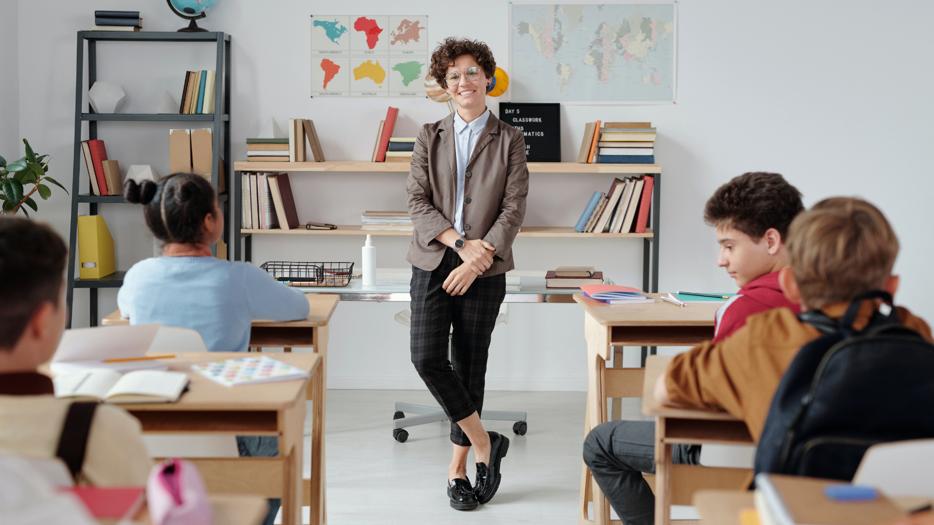 Zdjęcie przedstawia klasę szkolną. Na środku klasy, przodem do kadru stoi nauczycielka. Kobieta jest młoda, ma krótkie, kręcone włosy i nosi okulary. Ubrana jest w bluzkę z kołnierzykiem, żakiet i kraciaste spodnie. Obok nauczycielki znajduje się biurko, za nią regał z książkami i mapy na ścianie. Przed nauczycielką w ławkach, tyłem do kadru, siedzą uczniowie. W pierwszej ławce siedzi dziewczynka.