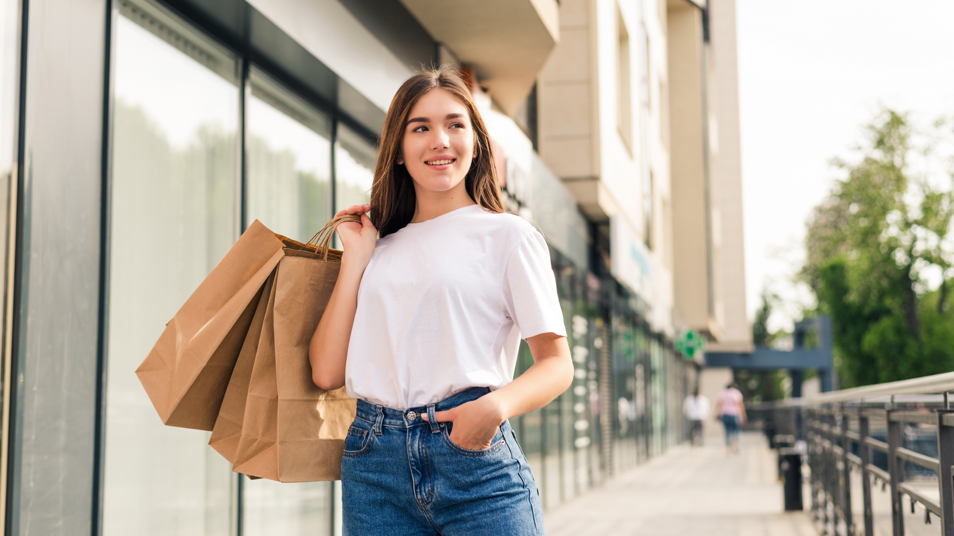 Zdjęcie przedstawia dziewczynę ubraną koszulkę i dżinsy. Na ramieniu trzyma dwie papierowe torby z zakupami. Dziewczyna znajduje się na zewnątrz obok budynku.