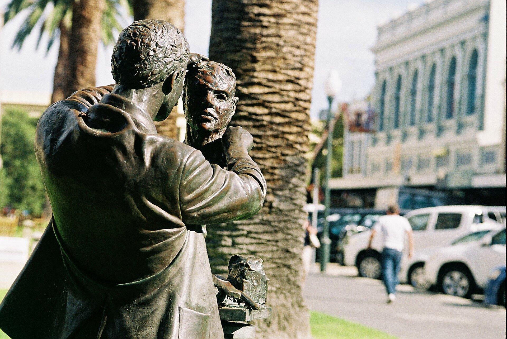 Zdjęcie przedstawia mosiężną rzeźbę. Jest na niej postać mężczyzny trzymającego w rękach głowę męską. Na drugim planie widać ulicę i rosnące przy niej palmy.