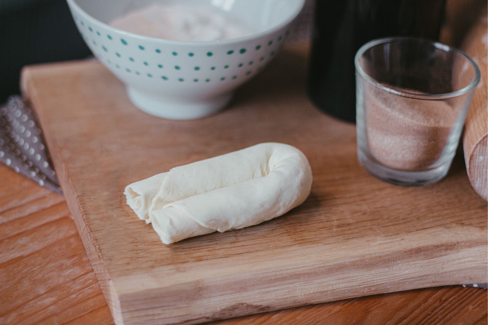Zdjęcie przedstawia krok piąty formowania serduszek. Ujęcie wykonane jest od góry pod kątem. W środkowej części grafiki widoczny jest zwinięty rulon ciasta złożony na pół i ułożony na drewnianej jasnobrązowej desce. Po prawej stronie od rulonu znajduje się przezroczysta szklanka wypełniona do połowy brązowym cukrem cynamonowym. Za nią dostrzec można czarny owalny kształt. Po prawo od szklanki widoczny jest fragment jasnobrązowego drewnianego wałka. Nad rulonem ciasta dostrzec można białą miseczkę w niebieskie małe kropki. Wszystkie przedmioty ustawione są na drewnianej desce, która leży na ciemnobrązowym drewnianym blacie. W lewym górnym rogu zdjęcia dostrzec można niewyraźny zarys ciemnej ścierki w białe kropki, która leży pod drewnianą deską.