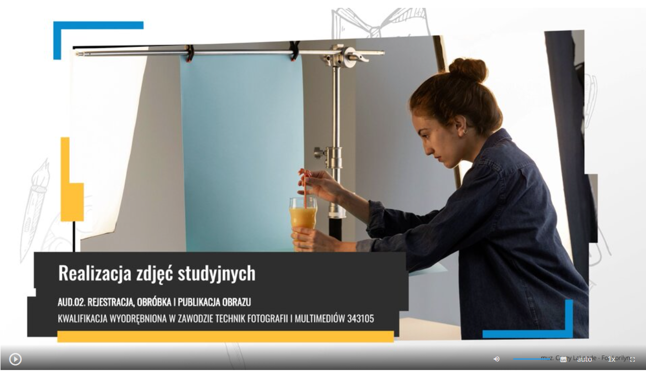 Zdjęcie przedstawia widok ekranu odtwarzania filmu instruktażowego. Po prawej widoczna jest postać kobiety skierowana w lewo, która trzyma w ręku szklankę z sokiem pomarańczowym z czerwoną słomką, które ustawia do zdjęcia. Powyżej widoczny jest l‑kształtny statyw, na którym powieszone jest szaro‑białe prostokątne tło. W lewym dolnym rogu widoczny jest napis z białych liter na czarnym tle – tytuł filmu Realizacja zdjęć studyjnych, oraz AUD.02. Rejestracja, obróbka i publikacja obrazu kwalifikacja wyodrębniona w zawodzie Fotograf 343101 i Technik fotografii i multimediów 343105.