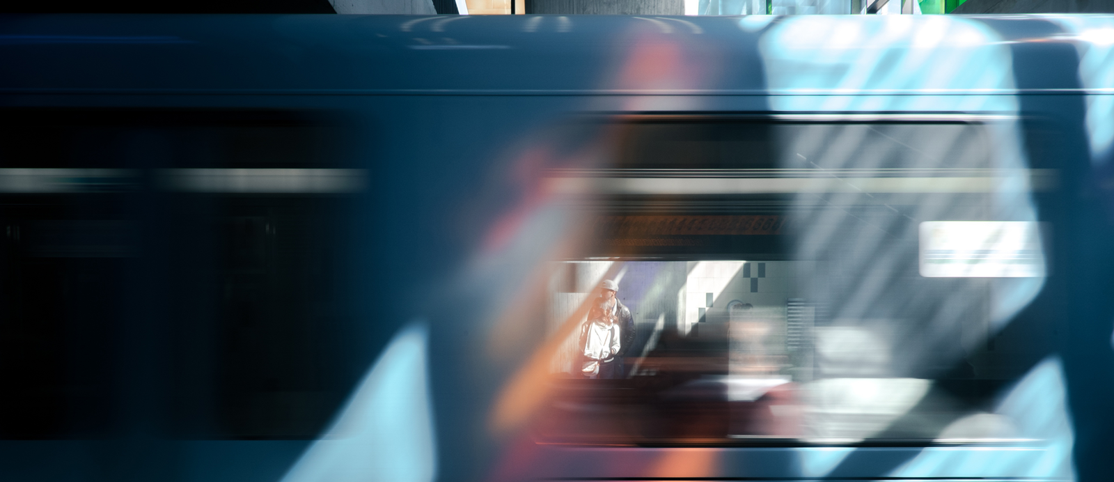 Zdjęcie jest nieostre, przedstawia zbliżenie przejeżdżającego pociągu. Widoczne są dwa okna, na jednym znajduje się nieczytelna prostokątna naklejka. W tle, przez jedno z okien, widać dwie osoby czekające na peronie oraz tablicę informacyjną – wyświetlany napis jest rozmazany.