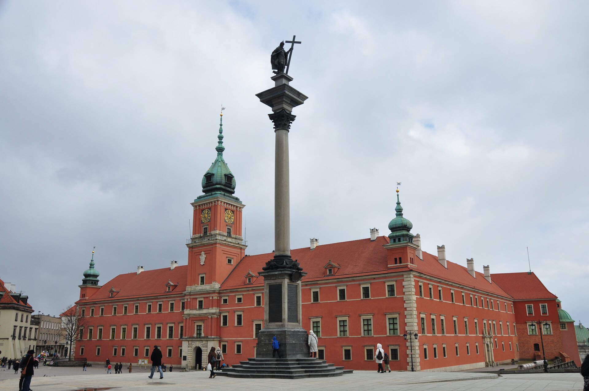 Na zdjęciu zachodnie i południowe skrzydło zamku królewskiego w Warszawie. Na pierwszym planie kolumna Zygmunta - wysoka kolumna na której stoi figura mężczyzny trzymającego krzyż. Pomnik stoi po środku placu. Za nim widoczny jest zamek - trzykondygnacyjny budynek. Po środku dłuższej z widocznych ścian, przylegającej do placu znajduje się wejście. Nad nim góruje wieża zegarowa zwieńczona kopułą. Widoczne są jeszcze dwie mniejsze wieże w narożnikach. Po placu przed zamkiem spacerują ludzie.