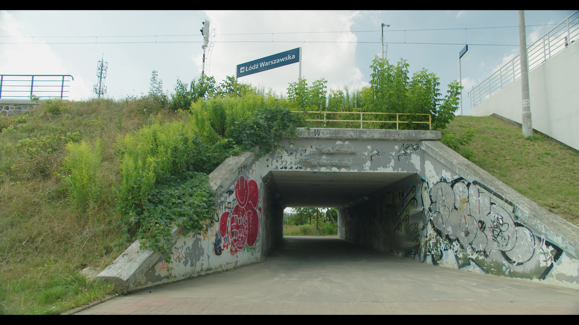 Ilustracja przedstawia przejście pod torami. Przez nasyp kolejowy obrośnięty trawą poprowadzone zostało przejście, którego wyjście znajduje się po drugiej stronie nasypu.
