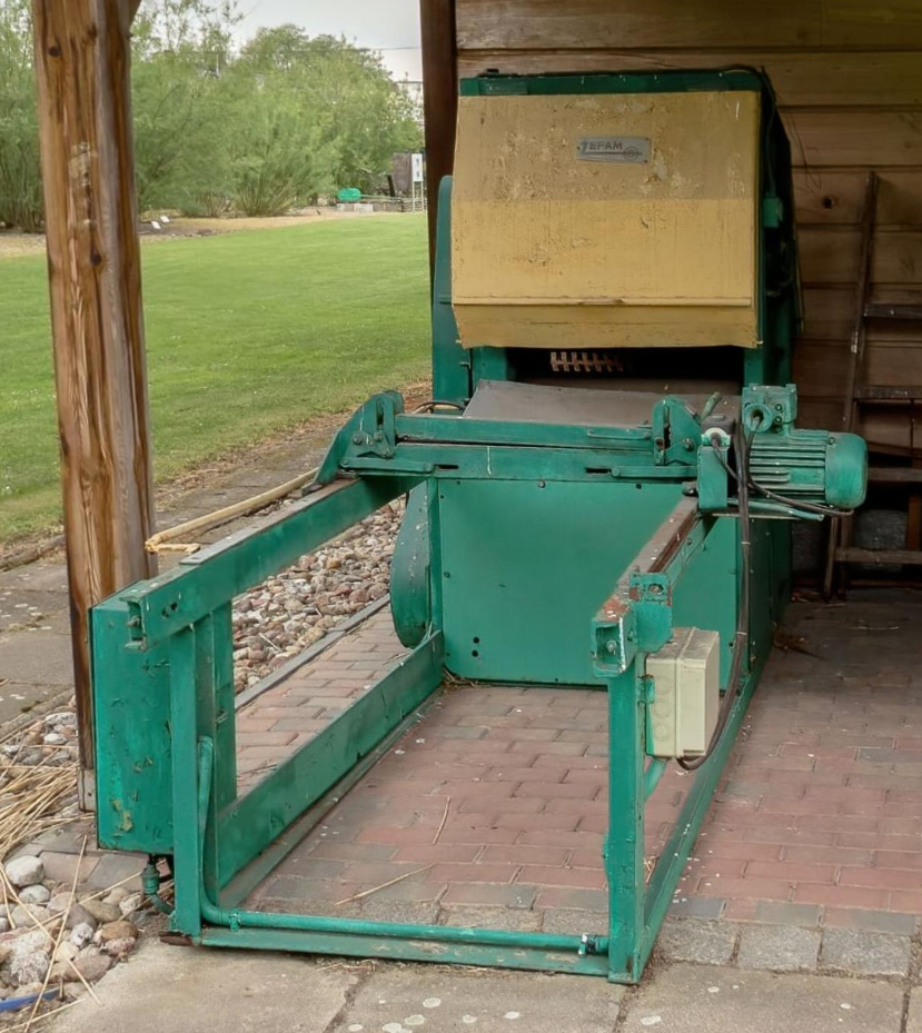 Zdjęcie przedstawia korowarkę stojącą na zewnątrz budynku. Widoczna jest metalowa obudowa oraz silnik. 
