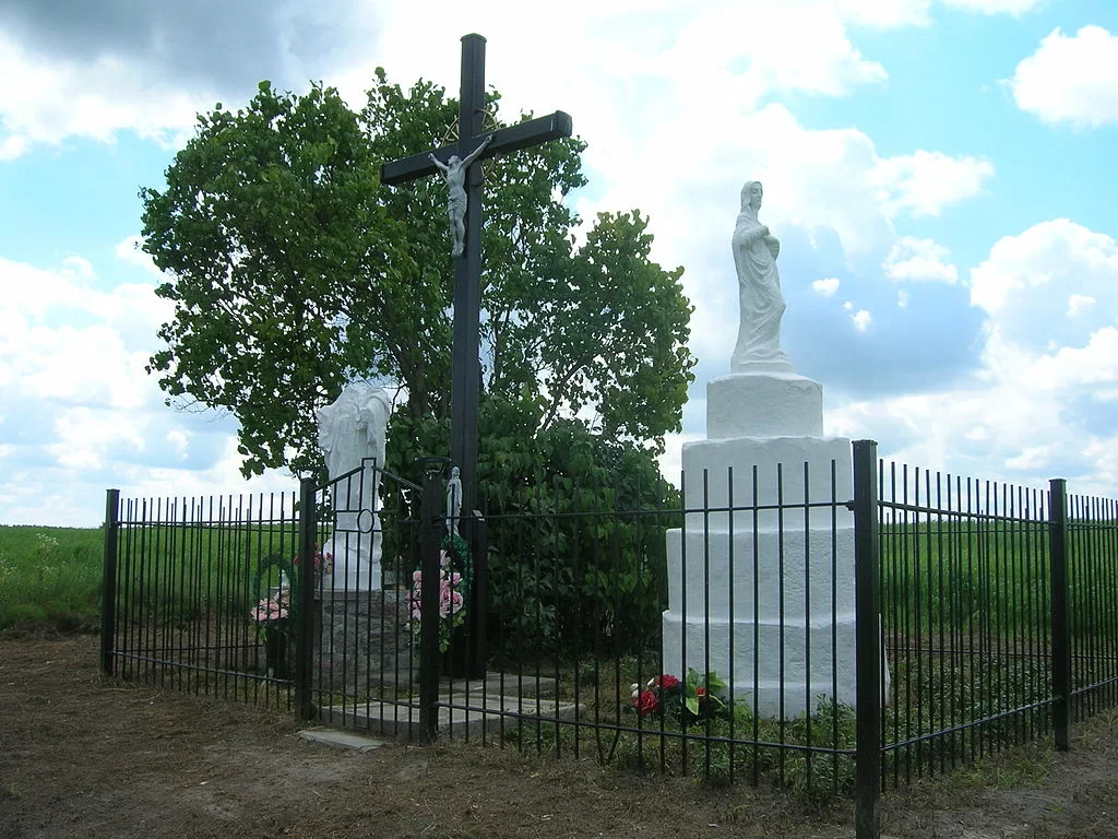 Zdjęcie przedstawia dwie białe figury Matki Boskiej oraz wysoki krzyż. Figura Matki Boskiej po lewej stronie nie ma głowy. Wszystkie trzy elementy są ogrodzone niewysokim, metalowym płotkiem. Wokół pola i łąki.