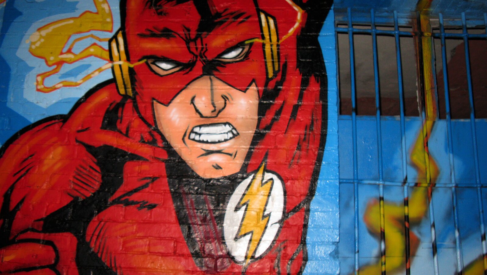 Fotografia barwna przedstawiająca fragment muralu z postacią bohatera komiksu Flash. Poczynając od lewej krawędzi, ponad połowę fotografii zajmuje pokryty farbą ceglany mur z ukazaną na błękitnym tle, od klatki piersiowej w górę, pochyloną ku przodowi postacią mężczyzny w czerwonym obcisłym kombinezonie z żółtą błyskawicą w okrągłym, białym polu na piersi. Z lewej strony w narożniku, podany ku przodowi i opuszczony niżej prawy bark, ukazuje wyraźnie zarysowane, napięte mięśnie. Wyżej na prawo i w górę głowa, której górna część nie została ujęta w kadrze.
