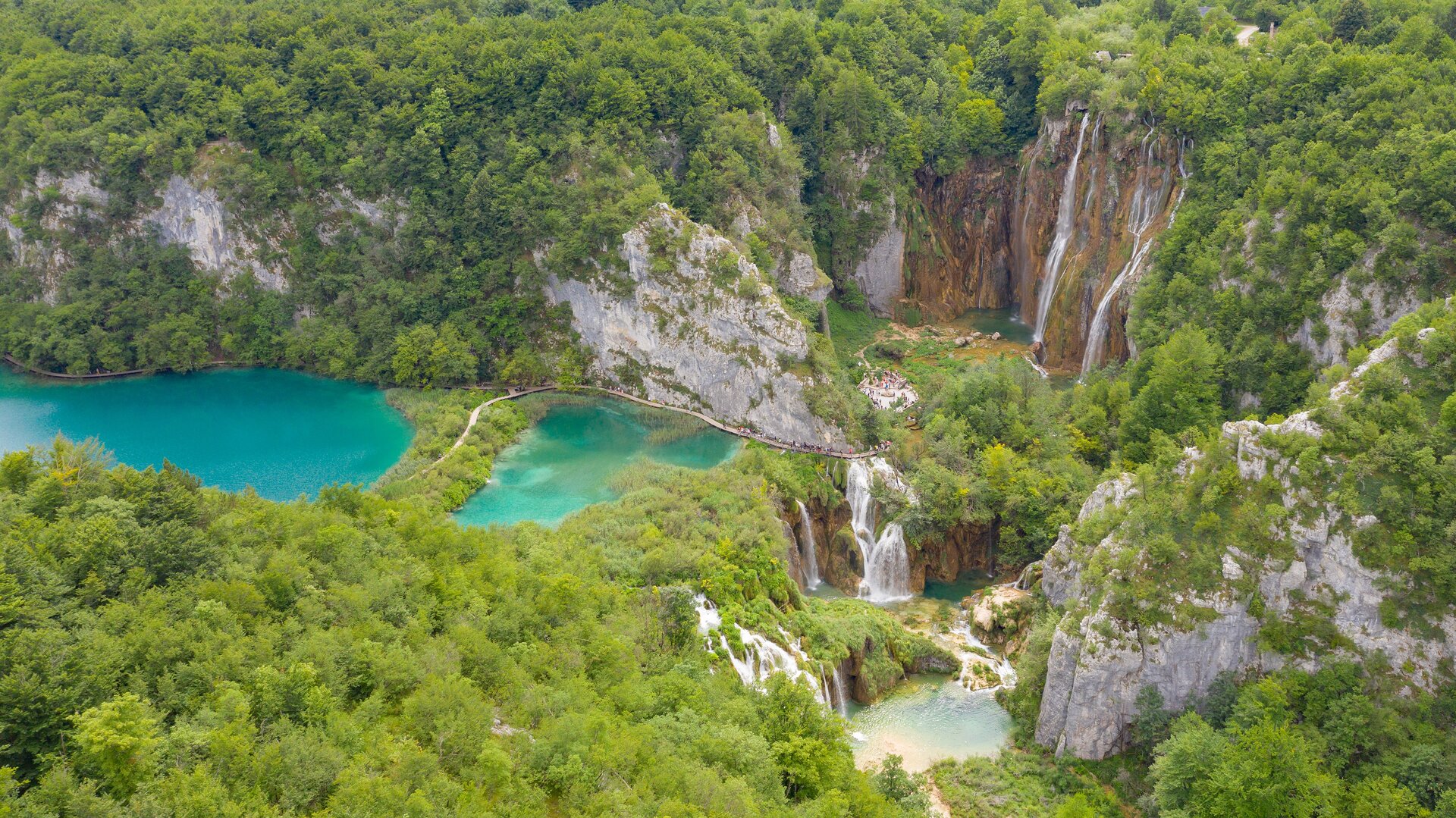 Na zdjęciu po lewej stronie bliżej środka widać dwa, nieduże, błękitne jeziora przedzielone naturalną granicą, na której znajduje się ścieżka. Po prawej stronie jeziora znajdują się ściany skalne koloru żółtobrązowego, na których spadająca woda tworzy wodospady. Takie same wodospady widać zarówno powyżej, jak i poniżej wodospadu środkowego. Całość otoczona jest skalnymi ścianami, porośniętymi na szczytach zielonymi drzewami.