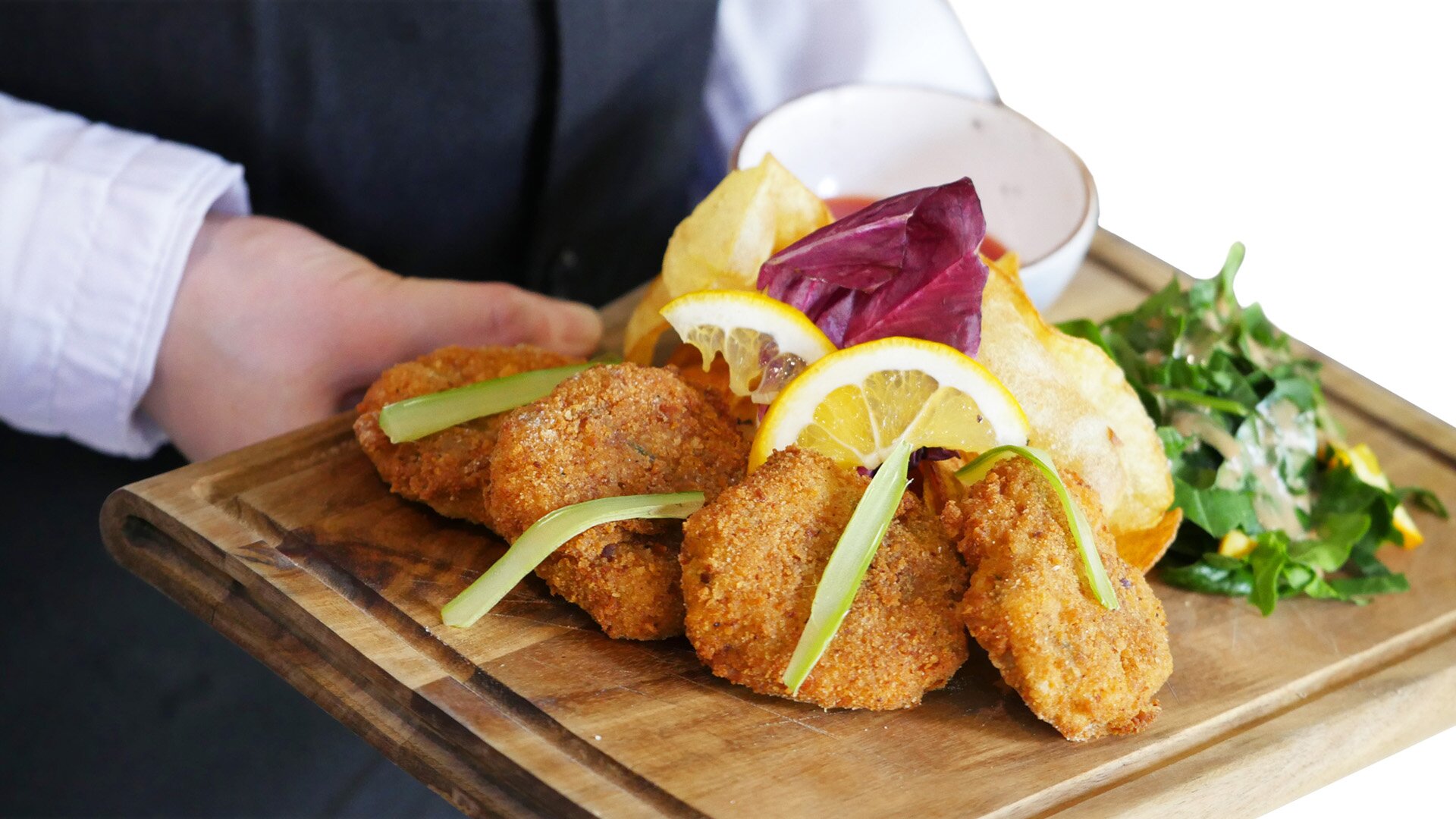 Na zdjęciu znajduje się trzymana przez kelnera potrawa. Na drewnianej desce widać cztery kawałki panierowanego kurczaka z cytryną, ziołami, ziemniakami i miseczką sosu.