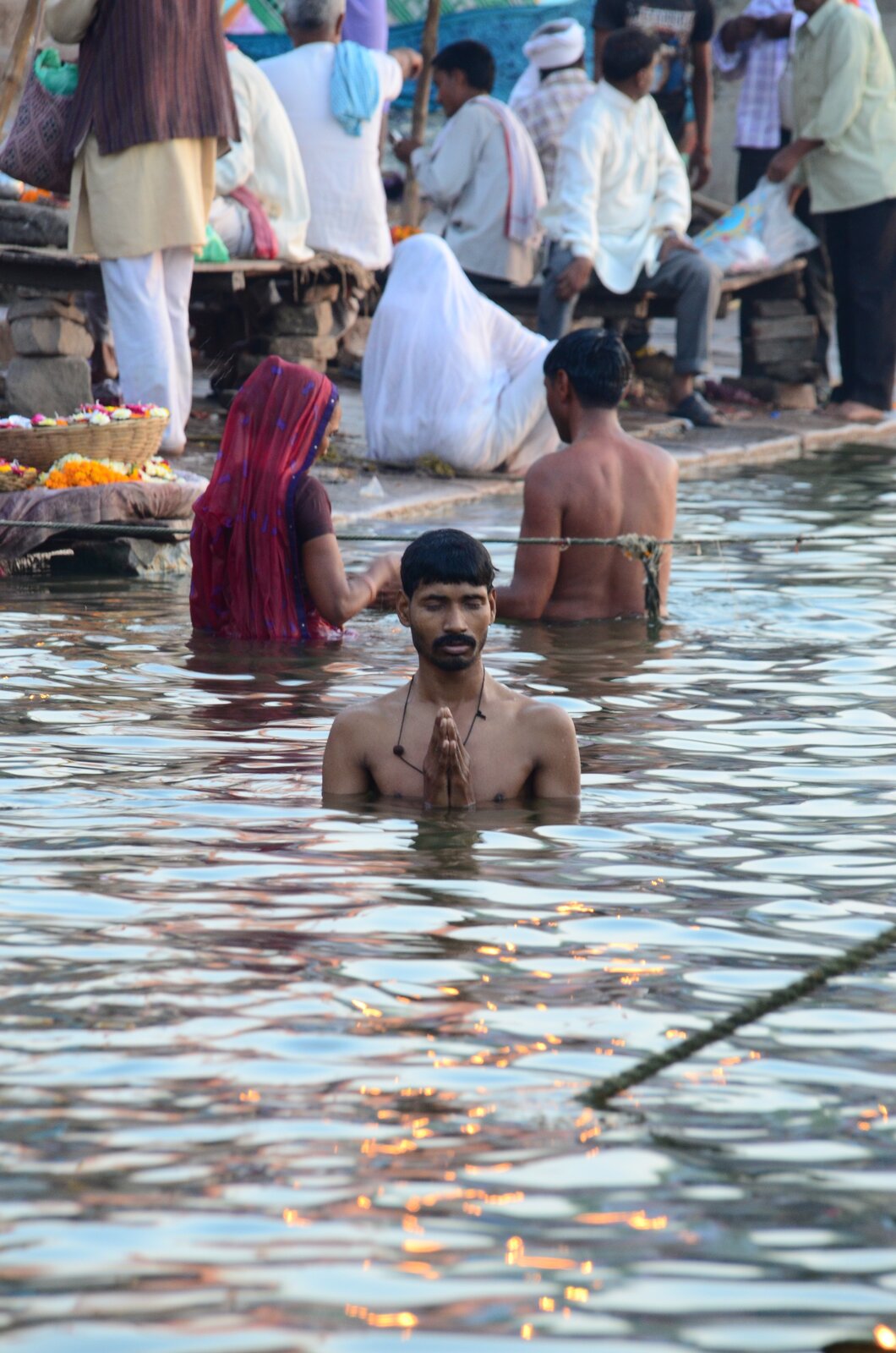 Zdjęcie przedstawia rzekę, w której blisko brzegu stoją trzy osoby. Na pierwszym planie jest mężczyzna z wąsami - Hindus. Ma zamknięte oczy i złożone do modlitwy dłonie. Woda sięga mu do klatki piersiowej. Tuż za nim w rzece stoi kobieta i mężczyzna, są odwróceni tyłem. Kobieta jest ubrana, ma tkaninę na głowie, która opada na plecy. Mężczyzna stojący obok niej ma nagie plecy. Na brzegu rzeki jest wiele osób - niektórzy siedzą, inni stoją.     