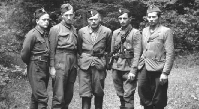 Kliknij, aby powiększyć Zdjęcie przedstawia stojących obok siebie pięciu żołnierzy. Mają na sobie niejednolite umundurowanie. Noszą koszule, marynarki albo wojskowe kurtki. Pozują na tle drzew.