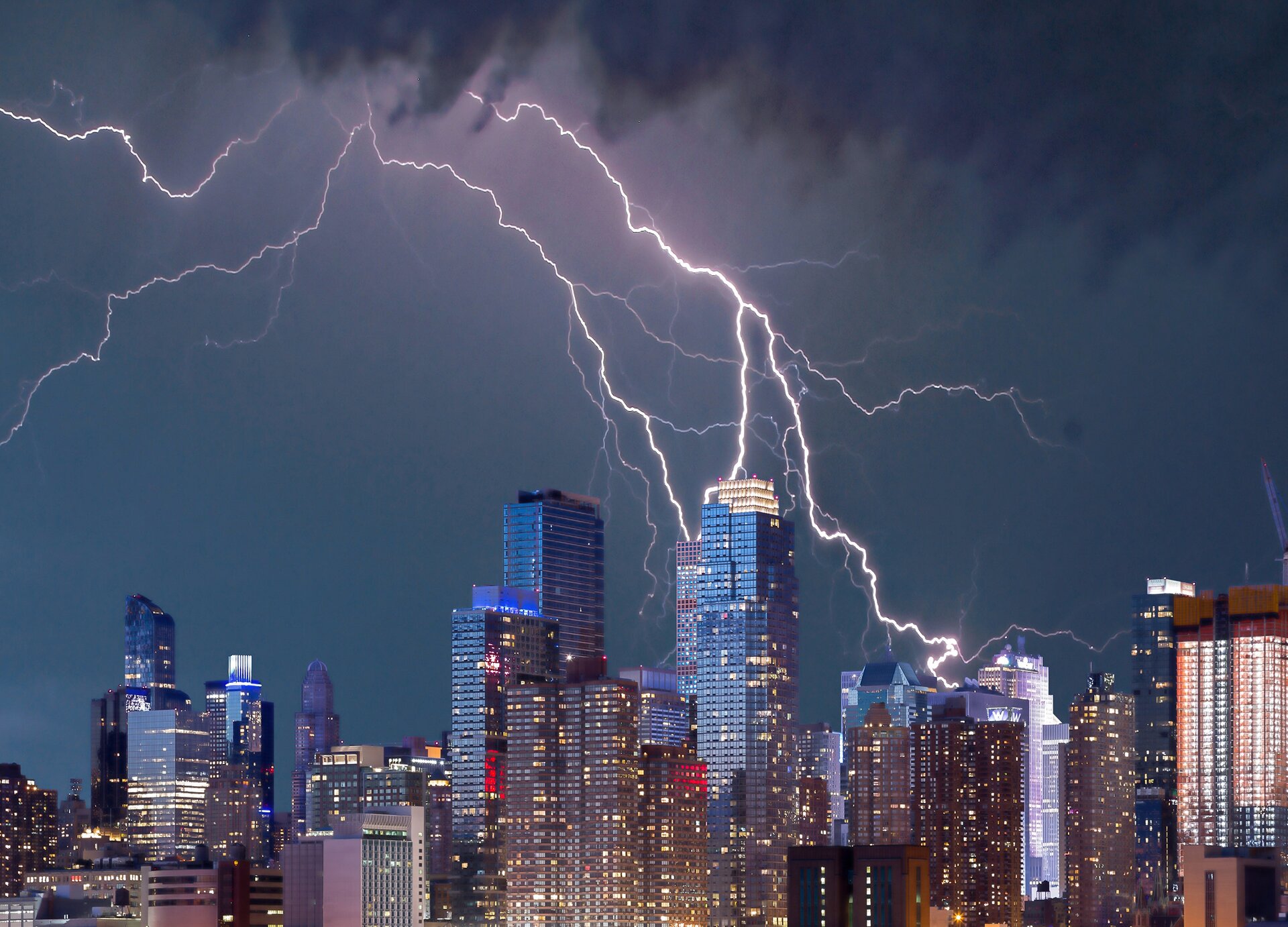 Zdjęcie przedstawia nocną panoramę dużego miasta z drapaczami chmur o różnej wysokości i formie architektonicznej. W wielu oknach budynków palą się światła. Niebo nad budynkami jest ciemnostalowe z białymi, rozgałęzionymi błyskawicami, które schodzą w kierunku najwyższych  budynków.